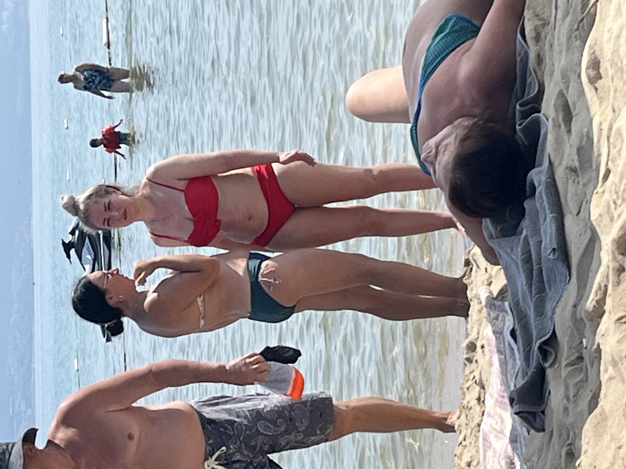 The image captures a sunny day at the beach in Nesebar, Bulgaria. The scene is a mix of people in the water and on the sand. In the foreground, partially on the sand and partially in the water, are two women in bikinis. One woman in a red bikini is lying on her side, with her arm resting on her hip. The other woman, wearing a teal bikini, is also lying down, facing away from the camera.
Further out in the water, two figures are visible. One appears to be a child in a red life vest, wading in the water. Behind them, another person in a patterned swimsuit is standing. The water is calm and reflects the bright sky.
On the sand, a person is lying down, with their legs stretched out. A blue and green patterned bikini bottom is visible, along with a towel draped over their legs. Another towel, light purple and white, is also on the sand. The sand appears light brown and slightly wet in some areas.
The overall impression is a relaxed summer day at the beach, with people enjoying the water and the sun. The time of day is likely midday, given the bright sunlight and the activities depicted. No visible text is present in the image.