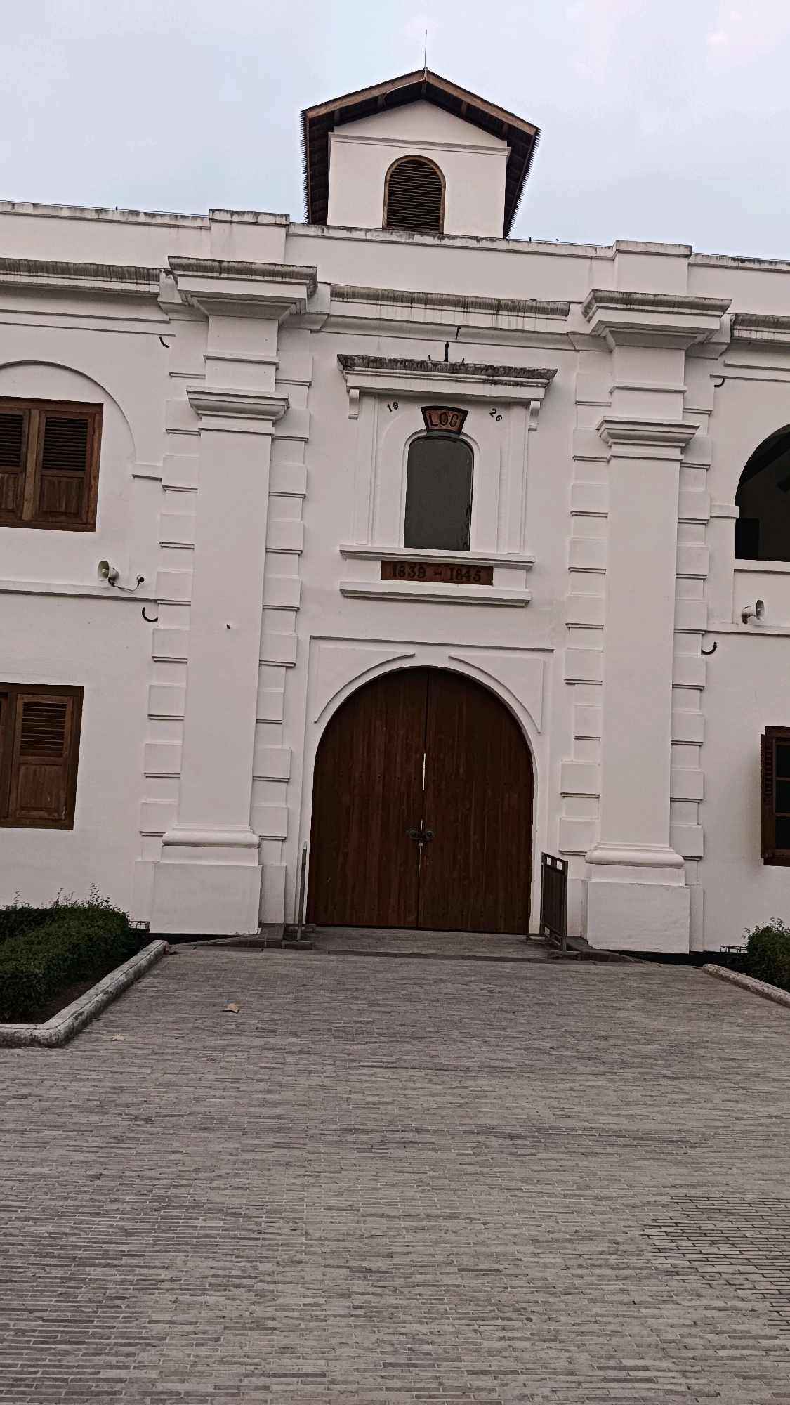 The image shows the front facade of a white colonial-style building with a large, arched wooden double door at its center. Above the door, a small, arched window is set into the wall, and above that, a decorative architectural element featuring the red inscription "1839-1845". The building has prominent vertical pilasters and horizontal cornices, typical of colonial architecture. Two smaller, similarly styled arched windows with wooden shutters are visible on the left side of the building. A small security camera is mounted on the wall to the left of the main entrance. The foreground is paved with grey brickwork, forming a sloping pathway leading up to the building. A neatly trimmed green hedge runs along the left side of the pathway. The sky above is overcast and grey. This building is located in Ngawi, Indonesia.