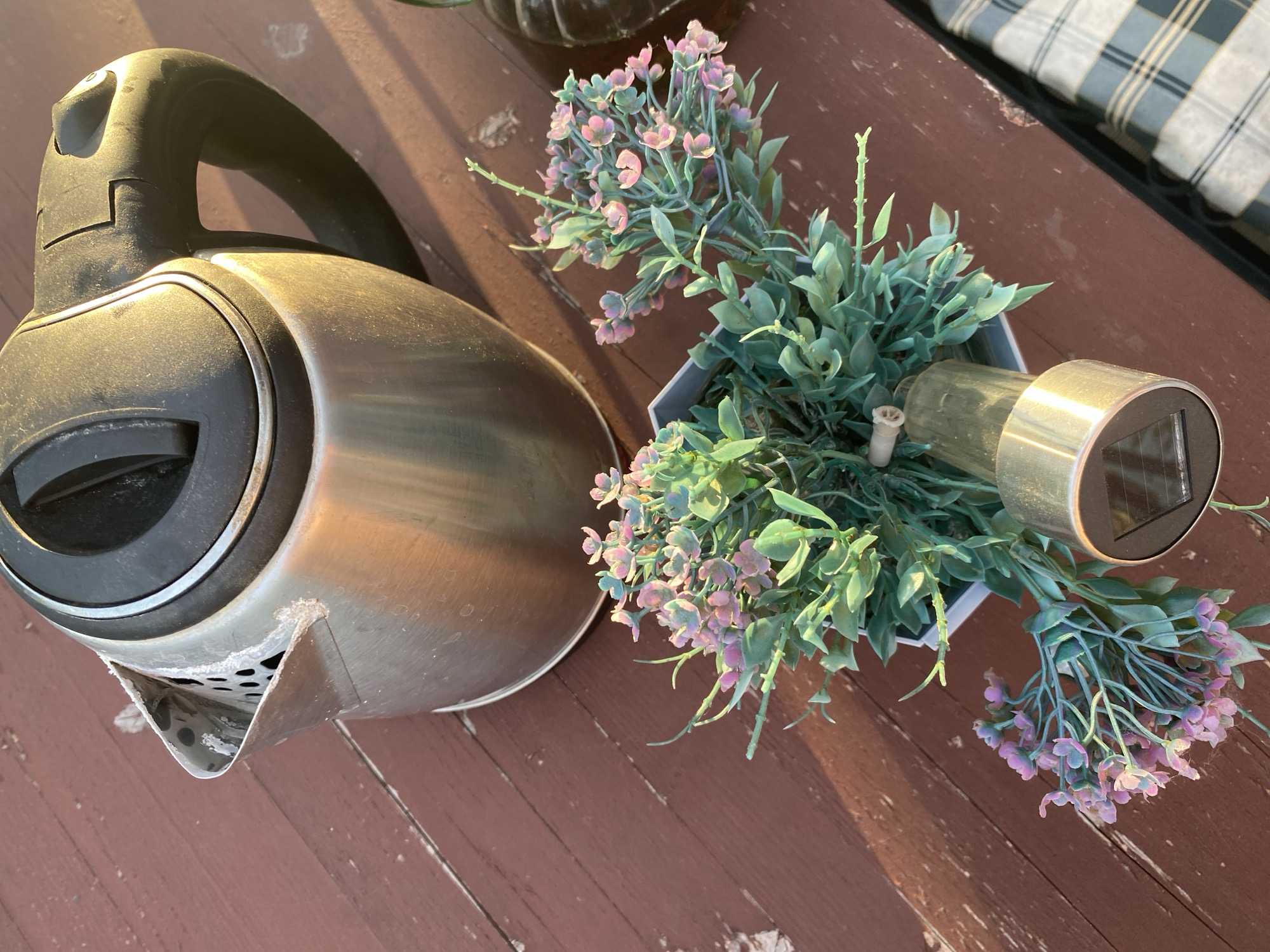 The image shows a close-up view of a stainless steel electric kettle with a black lid and handle placed on a weathered, reddish-brown wooden surface, likely a table. Next to the kettle is a small potted artificial plant with green foliage and clusters of pink and purple flowers. A solar-powered garden light is partially visible within the pot. The lighting suggests it is outdoors, possibly during sunset, casting long shadows. There are no people visible in the image. The setting appears to be a casual outdoor area, potentially a balcony, patio, or garden. The location is specified as Bol'shoe Zaborod'e, Russia, but the visual elements do not provide specific confirmation of this locale.