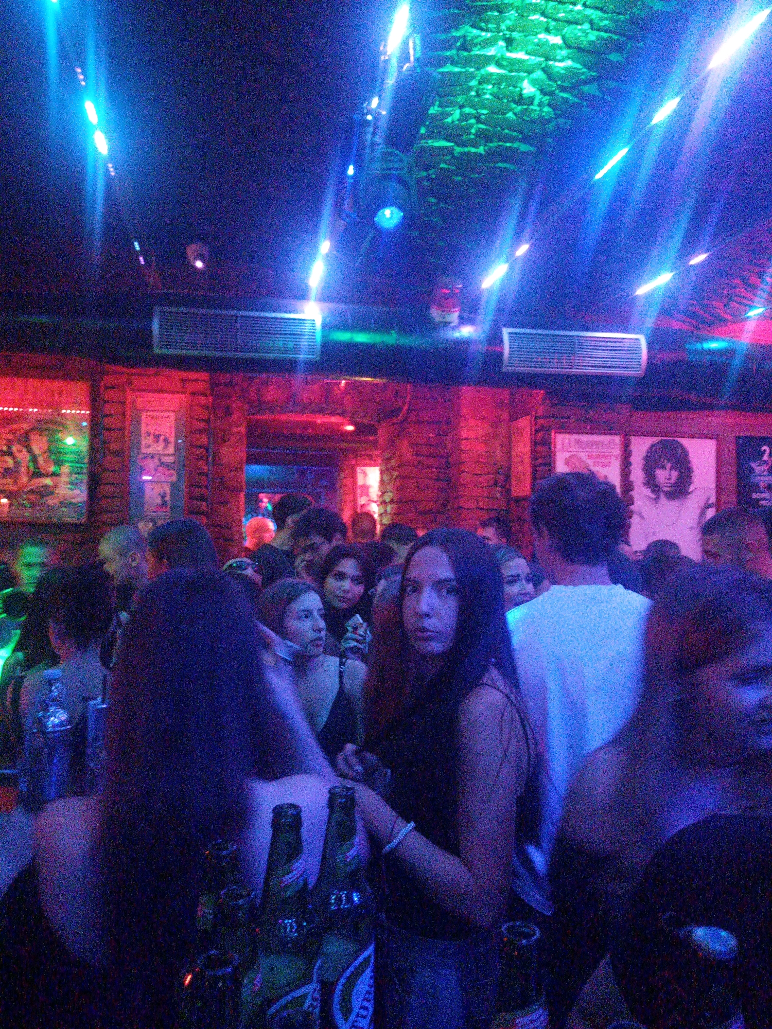 The image depicts an indoor scene, likely a nightclub or bar, with patrons gathered. The lighting is dim and colorful, with blue and green neon lights illuminating the space from above and a red glow emanating from the walls. The atmosphere appears lively, suggesting a social gathering. In the foreground, several individuals are visible, some in focus and others blurred. Two bottles of beer are prominently displayed on a surface in the lower right corner. The overall impression is one of a night out in a vibrant urban setting. It is not possible to determine the specific location within Blagoevgrad, Bulgaria, from the provided image.