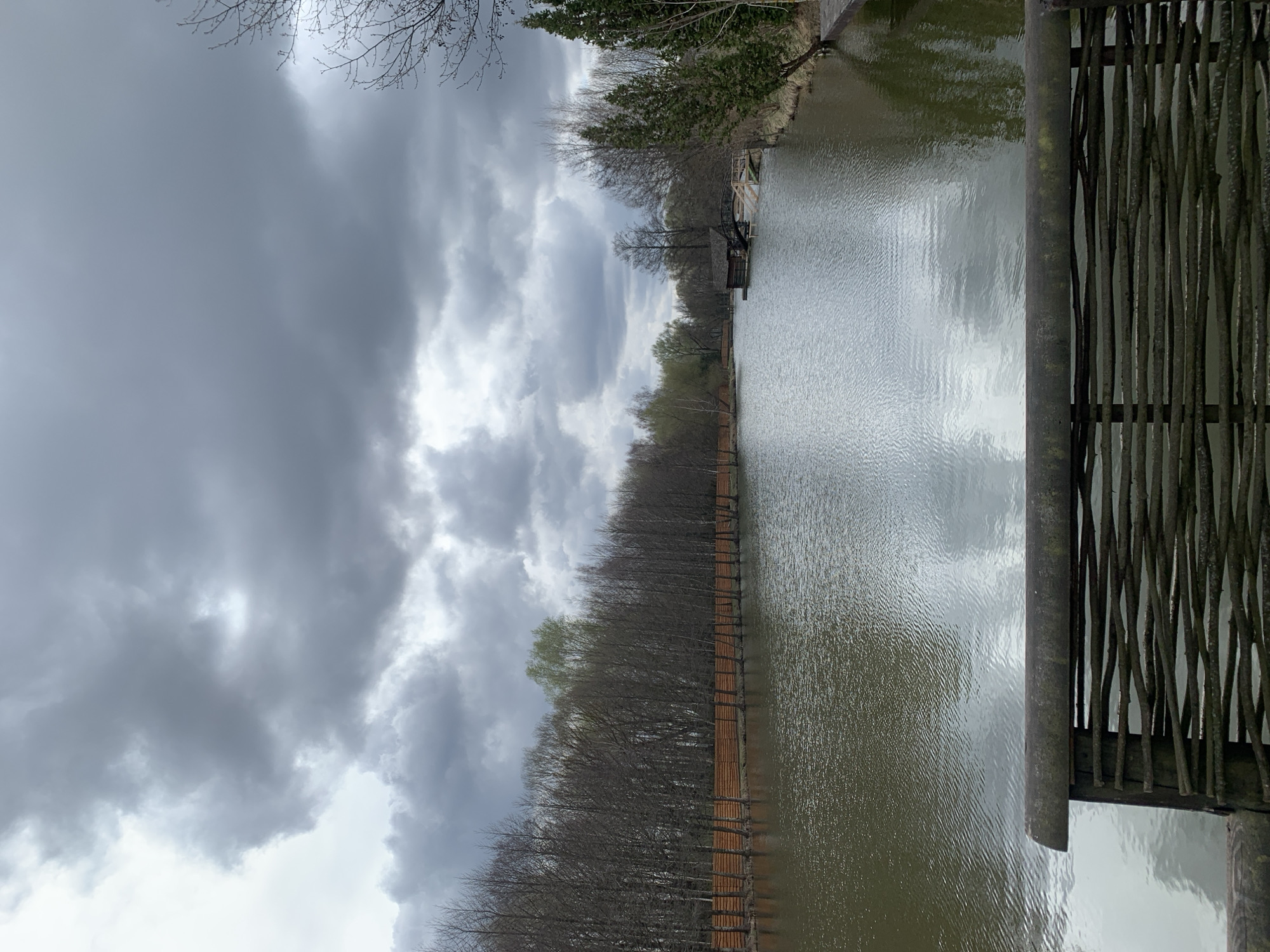 The image depicts a body of water next to a dense line of trees, under a cloudy sky. A rustic, wooden fence is visible on the right side of the frame. The scene is located in Капцёўскі сельскі Савет, Belarus. The water is reflecting the sky. A small structure is seen in the background. The trees appear to be bare or with minimal foliage.