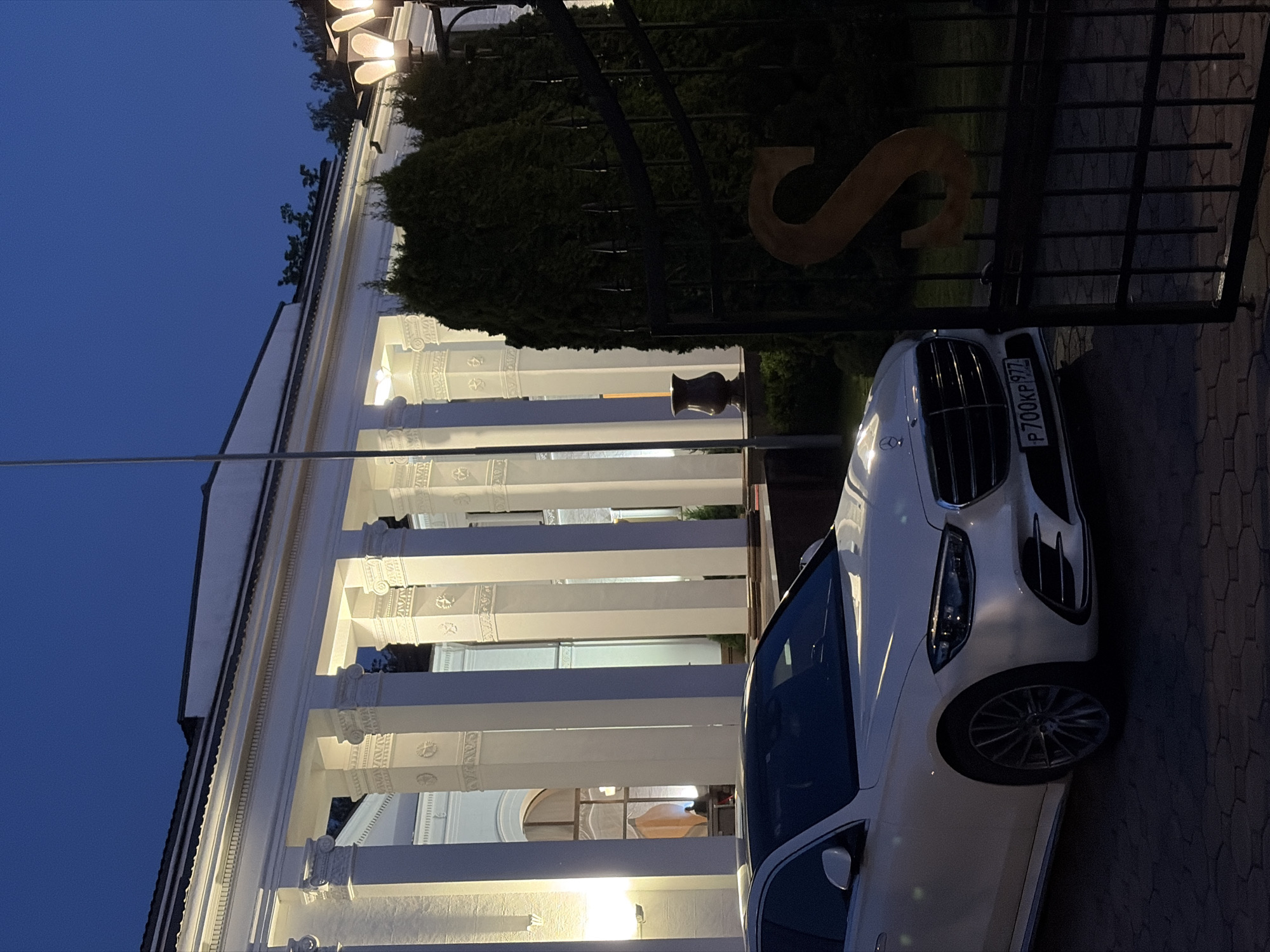 The image shows a white Mercedes-Benz parked in front of a building with white columns and a black wrought-iron gate. The car's license plate reads "P 700KP 977." A large, stylized "S" adorns the gate. The location is in an unidentified city. The sky is dark blue.