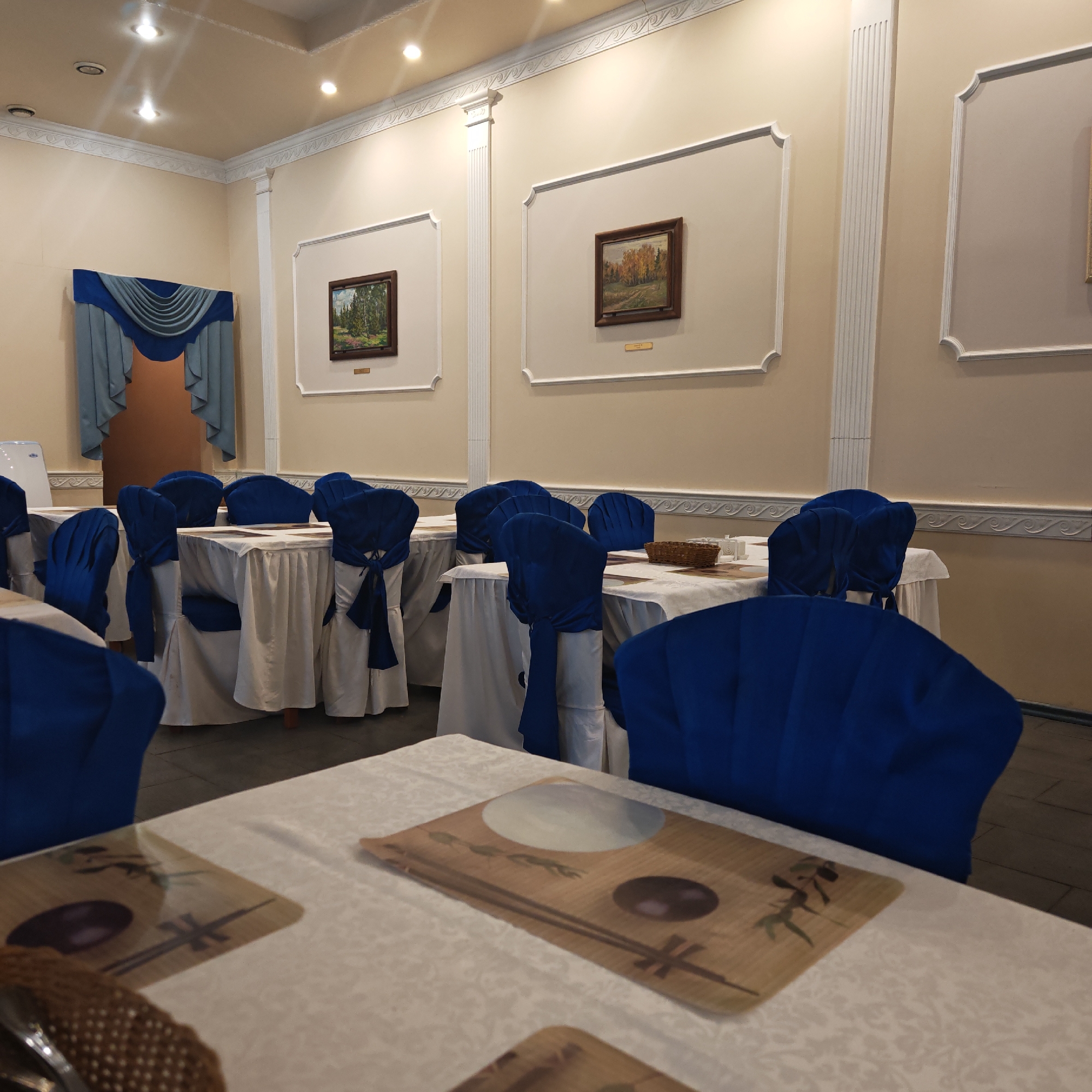 The image captures an empty dining hall or restaurant, meticulously set up for guests. The room features light-colored (beige/cream) walls adorned with ornate white decorative molding, including crown molding and fluted pilasters, creating a classical aesthetic. Several framed landscape paintings depicting forest scenes, one with green trees and another with autumn foliage, hang within these wall panels. Small, unreadable plaques are visible beneath each painting.
The ceiling is also light-colored and fitted with several recessed spotlights providing illumination. In the background, a doorway or archway is framed by elegant blue and grey draped curtains. A white appliance, possibly a water cooler, stands to the left of this archway.
The room is furnished with numerous tables draped in white tablecloths. Each chair is covered in a vibrant royal blue fabric, tied neatly with a bow at the back. On the tables closest to the foreground, brown placemats with Asian-inspired designs, featuring what appears to be a bowl, chopsticks, and green foliage elements, are visible. Some tables also have small woven baskets and white condiment containers. The floor is tiled or concrete, visible beneath the table coverings.
The setting appears formal and ready for an event or meal service, but no people are present. The lighting suggests an indoor evening setting or an area reliant on artificial light. The overall atmosphere is quiet and orderly.