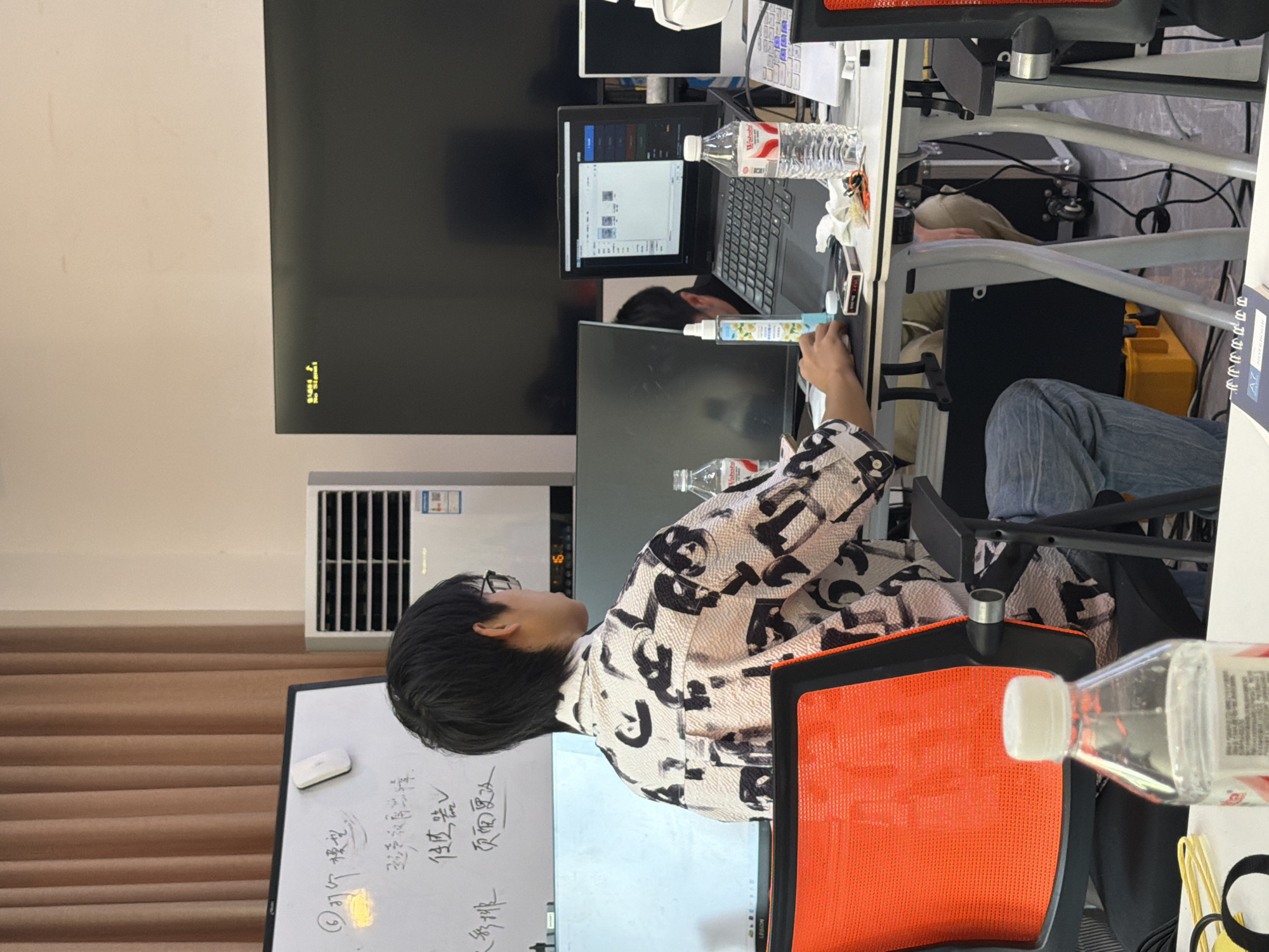 The image depicts an indoor scene, likely an office or classroom environment, with a focus on a young person seated at a desk. The person, appearing to be male with short dark hair and glasses, is wearing a patterned shirt and jeans. They are looking towards a laptop computer which is open on the desk. The desk also holds other items including a water bottle, a spray bottle, and a pack of cards. In the background, there are multiple computer monitors, a whiteboard with writing on it, an air conditioner unit, and a large black screen. The overall impression is one of a workspace or study area. The location is identified as Jiangjin, China.