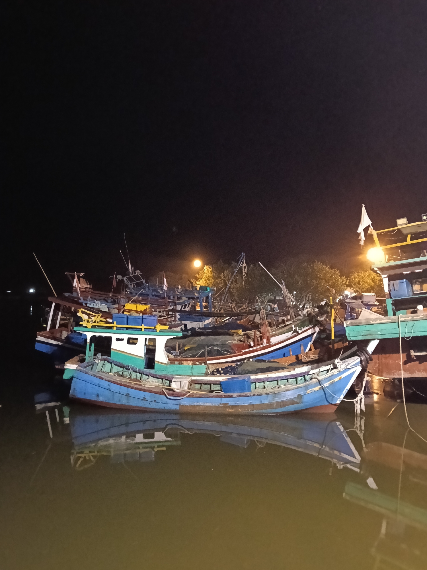 The image presents a nighttime scene of several fishing boats moored in a calm harbor or river, likely in Lhokseumawe, Indonesia. The sky is dark, indicating a clear night, with artificial lights providing illumination.
The main subjects are numerous wooden fishing boats, densely packed together. The boat closest to the viewer is painted a vibrant blue on its hull with a lighter blue, white, and green cabin structure. Its detailed reflection, including parts of its hull and cabin, is visible on the still, dark green-brown water. Other boats behind it display a mix of colors, including red, brown, white, and various shades of blue and green. Their masts, rigging, and various equipment are silhouetted against the night sky and the ambient light.
One boat in the middle distance appears to have the word "CRiSSA" or a similar string of letters faintly visible in white on its blue hull.
Bright, warm-toned lights, possibly from streetlights or dock lights, illuminate the scene, casting an orange and yellow glow on some boats and the surrounding trees in the background. These lights create prominent reflections on the water, adding to the visual depth. The trees, partially visible behind the boats, suggest a nearby shoreline.
There are no people visible, implying the boats are at rest. The overall scene conveys a peaceful and quiet atmosphere of a working harbor at night.