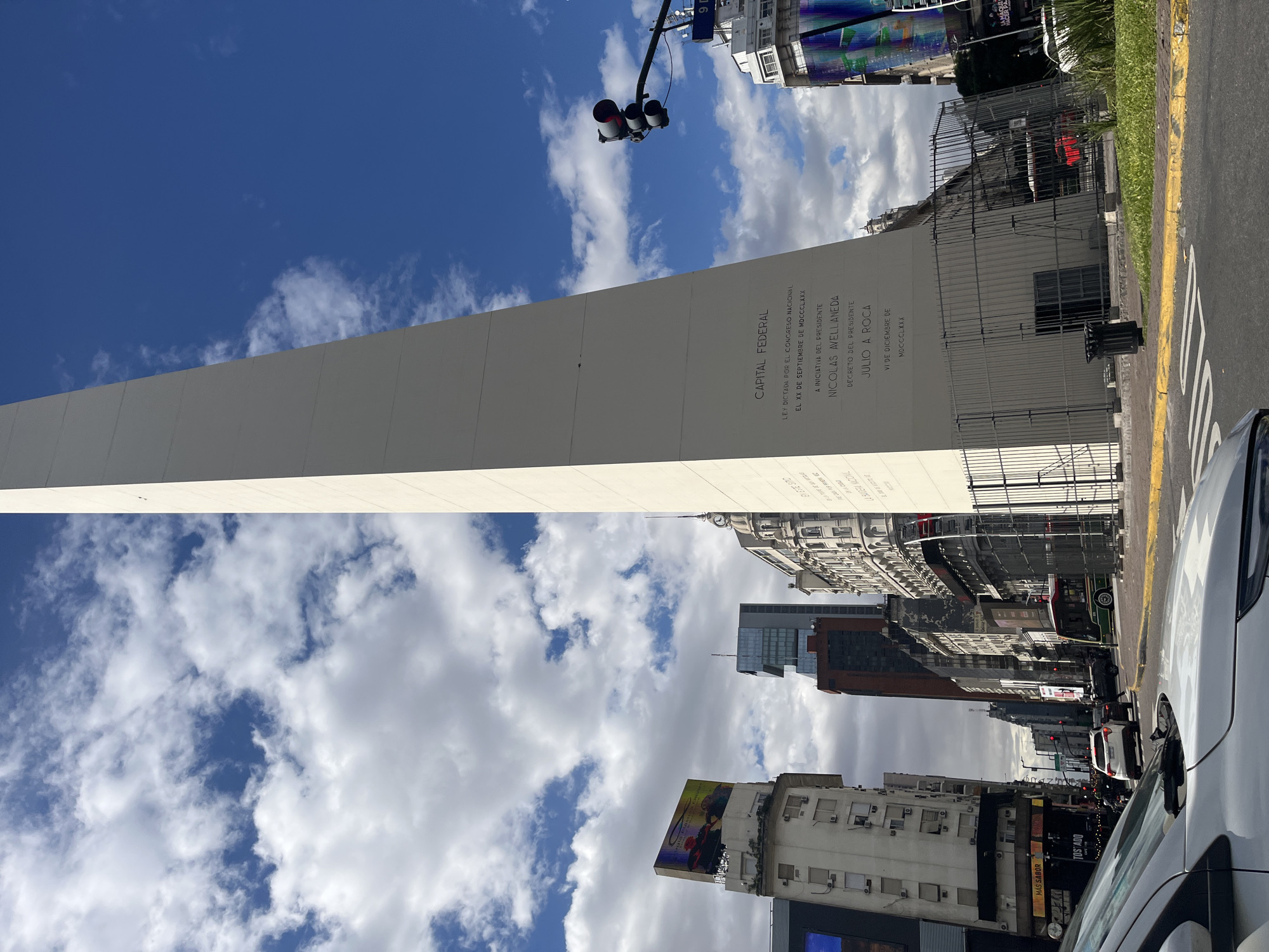 The image shows the Obelisco, a historical monument in downtown Buenos Aires, Argentina. The obelisk, a tall, white, quadrangular spire, rises prominently against a partly cloudy sky. The monument is inscribed with text including "Capital Federal," the name of the city, and names of presidents. Surrounding the monument are city buildings, advertising signs, and a traffic signal. A street with cars, including a white vehicle in the foreground, runs alongside the obelisk. The picture has been taken from a low angle, giving prominence to the monument.