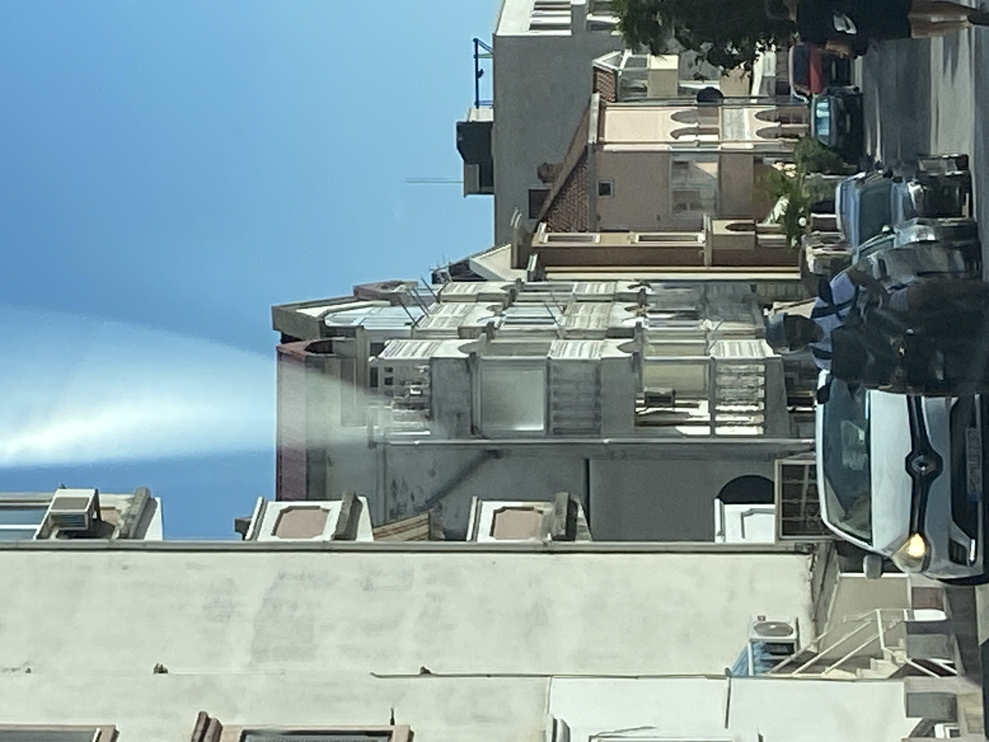 In Budva, Montenegro, on a sunlit day, a street scene unfolds with residential buildings lining a narrow road. A white Renault car is parked in the foreground, facing the viewer, with its headlights illuminated. To the left of the car, a person on a scooter, wearing a helmet and a white t-shirt, is visible. Further down the street, several other cars are parked and moving. The buildings, with their distinct architectural styles, feature balconies and numerous windows, suggesting a residential area. The sky above is a clear, bright blue.