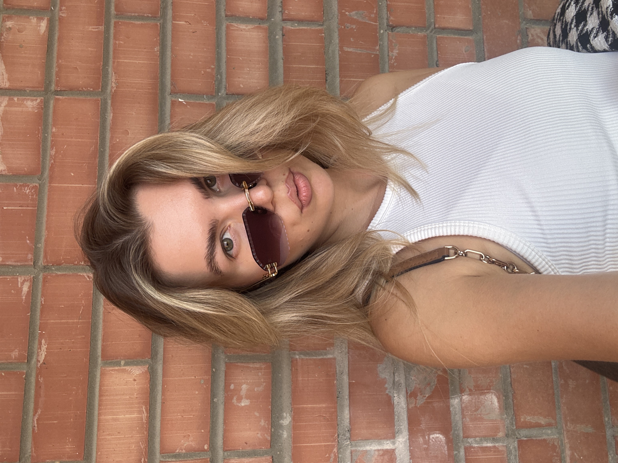 A young woman with blonde hair and sunglasses poses against a brick wall. She is wearing a white ribbed tank top and her arm is visible. The brick wall appears to be the exterior of a building, possibly in an urban setting. The lighting suggests it is daytime.