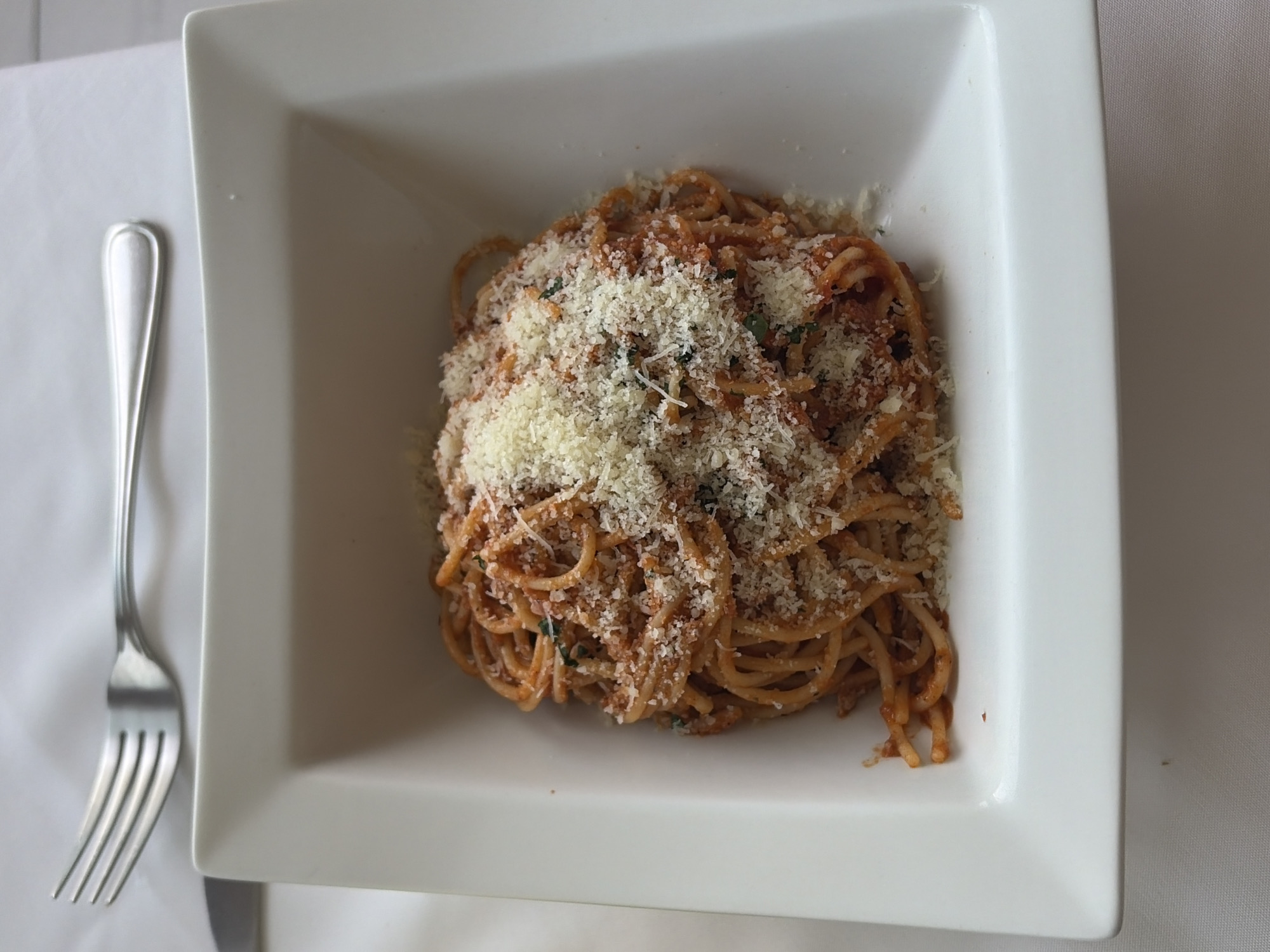 A top-down, medium shot shows a square, white porcelain bowl filled with spaghetti with tomato sauce and a generous topping of grated cheese, likely parmesan. To the left of the bowl, on a white tablecloth, is a silver fork, positioned parallel to the bowl. The scene appears to be in a restaurant setting. There is no indication of the location being in the city of Puebla, Mexico.