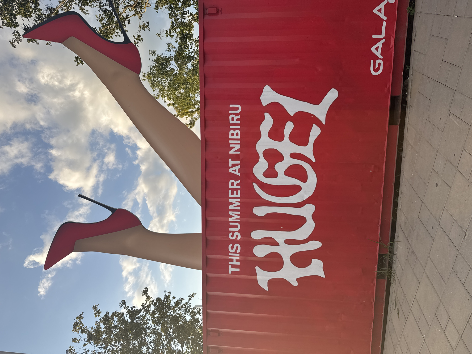 The image shows a large red shipping container in Costinești, Romania, with a giant sculpture of two legs wearing red high heels sticking out of the top, against a cloudy blue sky. The container has white text on it, including "THIS SUMMER AT NIBIRU" and a large, stylized white word that appears to be "HUGE!". In the lower right corner of the container, the word "GALA" is visible in white text. The container is situated on a paved sidewalk, with some patches of grass growing along its base. Trees are visible in the background. The perspective is from ground level, looking up at the sculpture and the container.