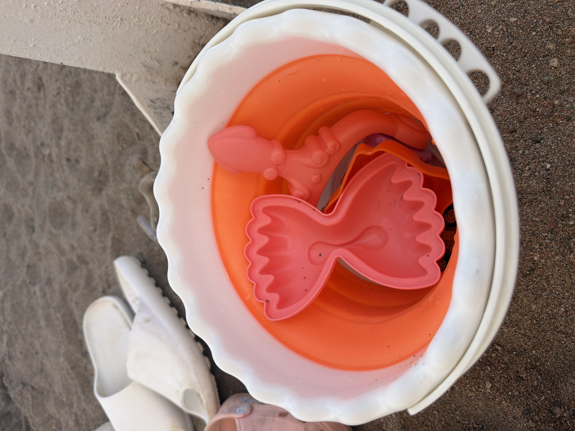 The image depicts a close-up view of a white bucket containing orange plastic sand molds, including a crab and a squid. The bucket is placed on a sandy surface, likely a beach, with a pair of white slides visible in the background. The lighting suggests it is daytime. The scene is static and does not depict any ongoing activity or individuals. No specific location in Choktal, Kyrgyzstan, is identifiable from the image alone.