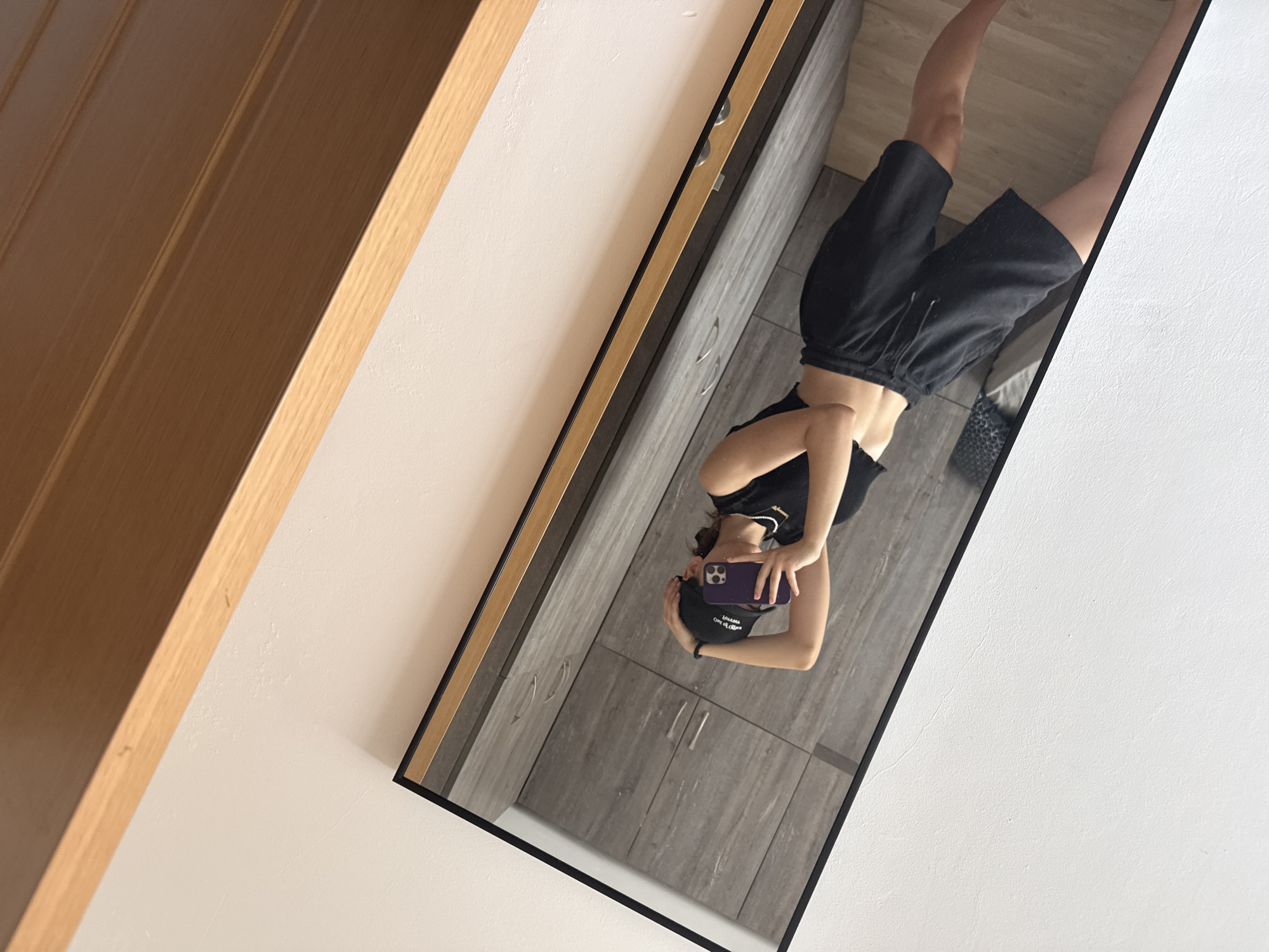 A medium shot depicts a mirror reflecting a person taking a selfie. The individual, dressed in black shorts, a black top, and a black baseball cap, holds a purple cell phone. The reflection also reveals grey, wood-grain patterned cabinets with silver handles. The mirror is framed in black. The surrounding walls are white, with a wooden doorway visible on the left side of the frame. No information suggests the location is Plovdiv, Bulgaria.