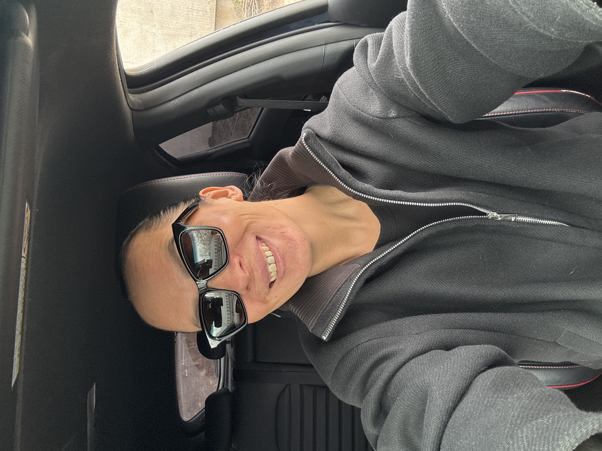 The image shows a person in a car, likely taken in Chaikovsky, Russia. The person is wearing sunglasses and a dark-colored jacket with a zipper. The interior of the car is predominantly black. Reflected in the sunglasses are building facades, which, combined with the prompt's geographical specification, suggest the photo was taken in an urban area of Chaikovsky. The individual is smiling slightly and appears to be taking a selfie. The background includes the car's interior and a glimpse of the outside environment through the windows.