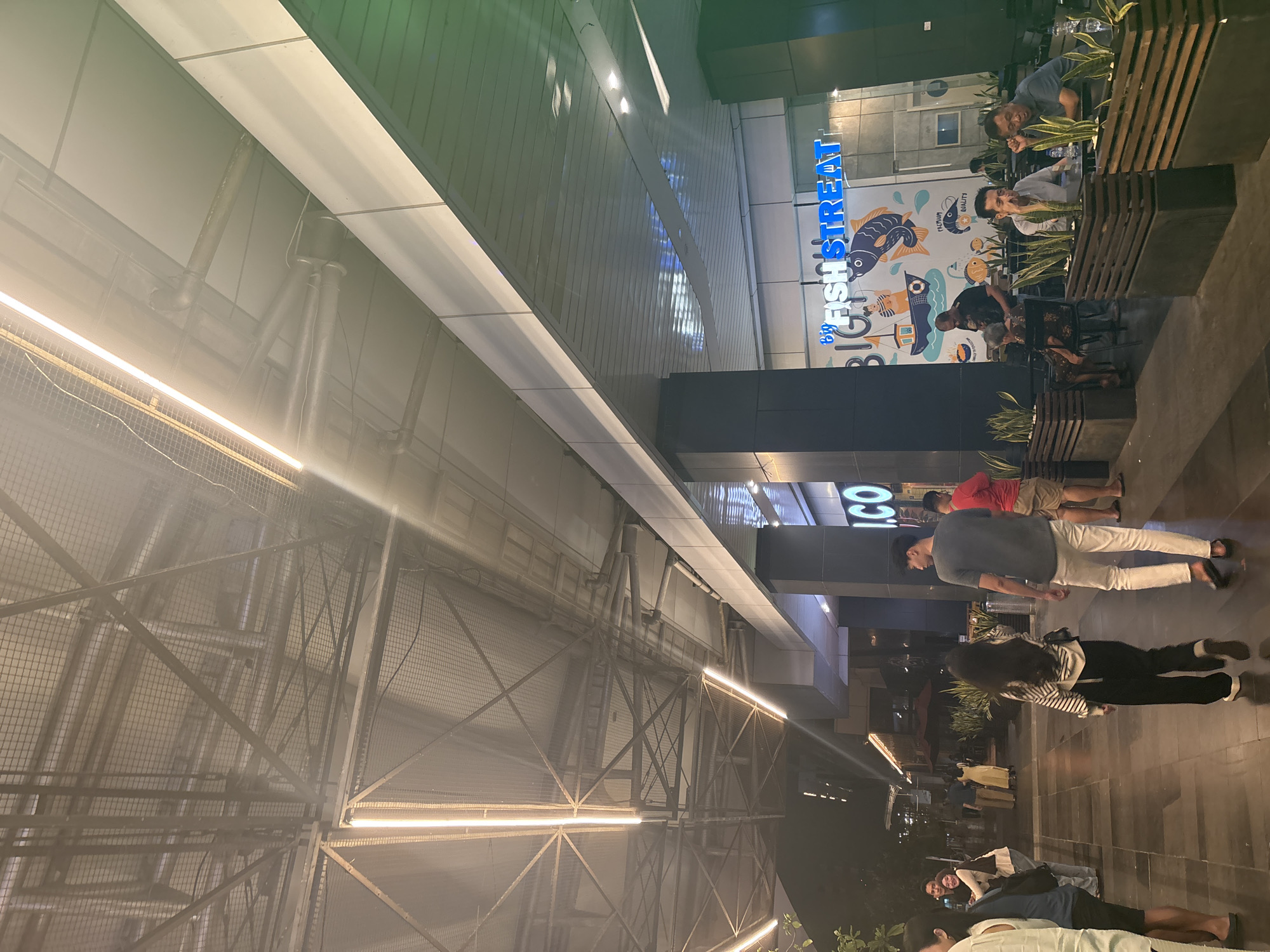 The image shows an outdoor dining area in North Jakarta, Indonesia, outside a restaurant called "Big Fish Street". Patrons are seated at tables, while others stroll along a paved walkway. The building structure incorporates exposed metal framework illuminated by strip lighting.