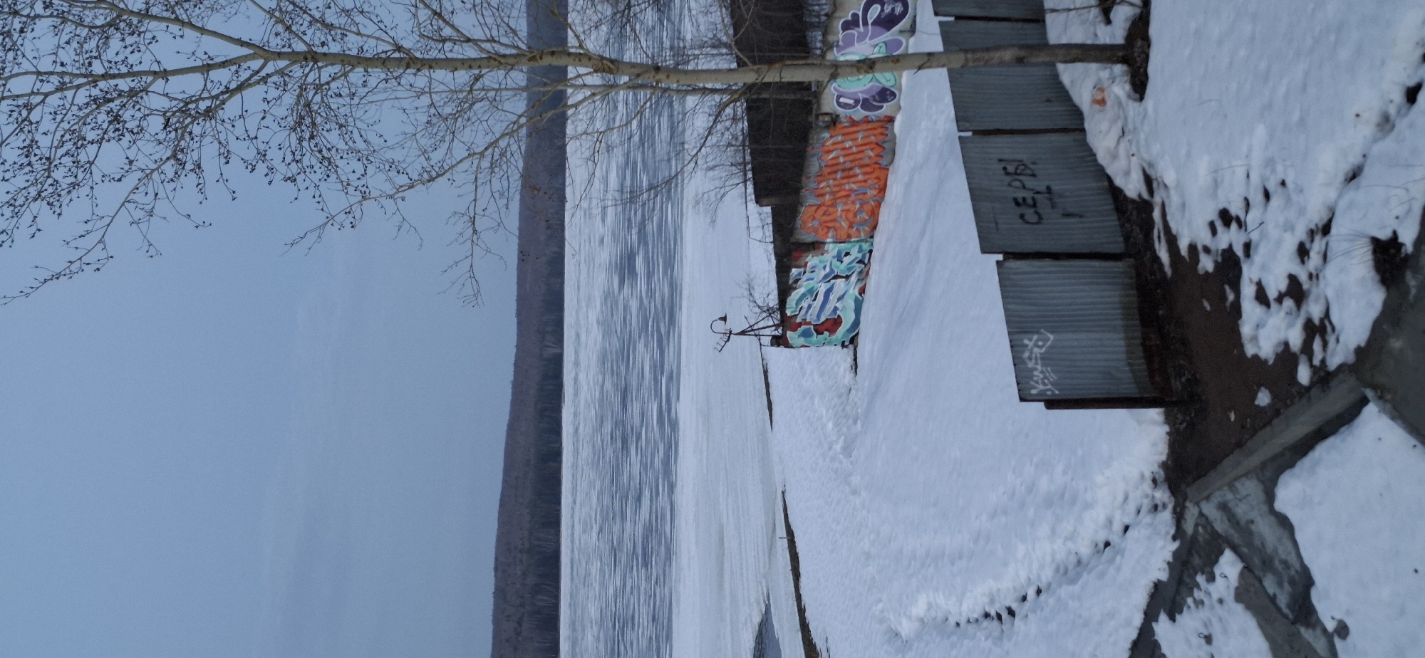 The image shows a winter scene near the Kama River in Chaikovsky, Russia. The river is partially frozen. A snow-covered embankment runs along the riverbank, featuring a concrete walkway and metal benches. Graffiti covers a concrete wall along the walkway. A bare tree branch extends into the frame, contrasting with the blue sky and the river. A lamp post is visible near the graffiti-covered wall.