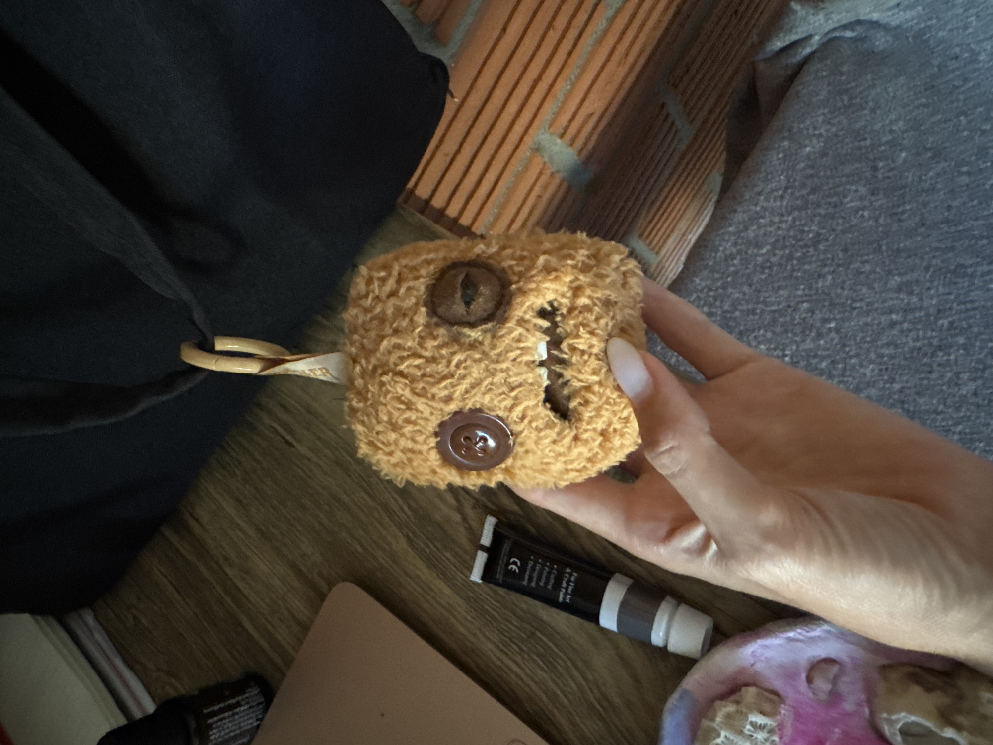 A handmade, fuzzy, orange creature with two buttons for eyes and a sewn mouth is held by a person's hand. The creature has a small loop attached to its head. In the background, a dark fabric bag, a wooden surface, a tube of cosmetic cream, and a brick wall are visible. There is no indication of the image being taken in Baan Nok Sai, Thailand.