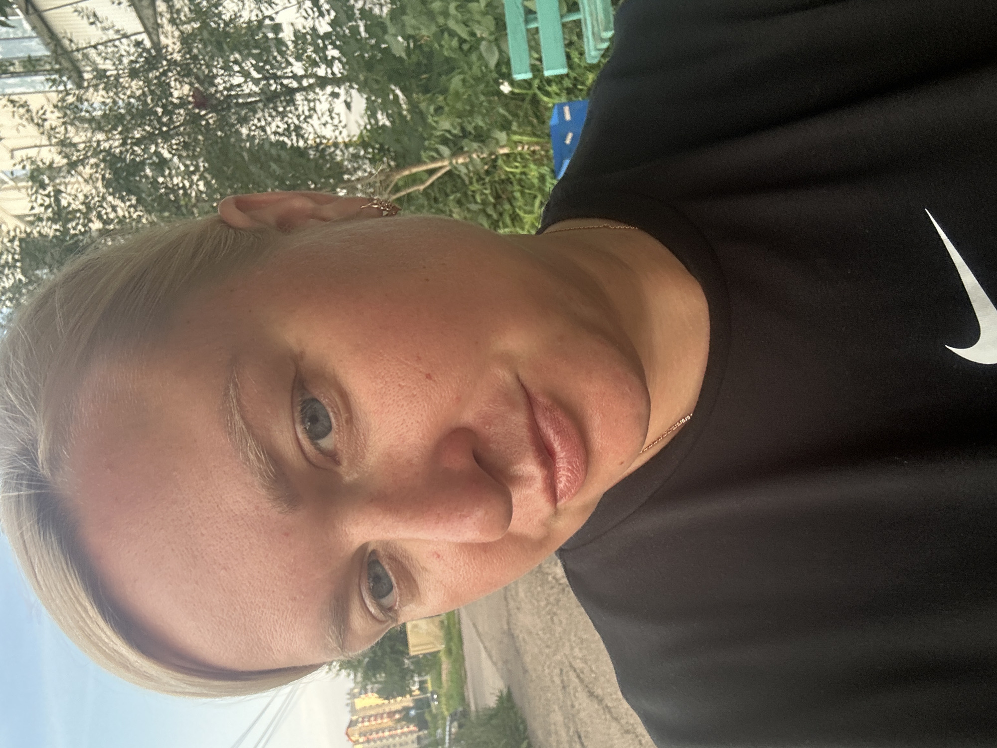 The image shows a close-up, selfie-style view of a woman with blonde hair and blue eyes. She is wearing a black t-shirt with a white Nike logo visible on the chest. Her face is slightly tilted, and she is looking towards the camera. Behind her, a blurry background includes a building, trees, a paved area that appears to be a sidewalk or road, and a glimpse of a green park bench and a blue trash bin. The lighting suggests it is daytime, possibly late afternoon or early evening. The setting appears to be outdoors in an urban or suburban environment. While specific details are difficult to ascertain from the visual alone, the presence of residential buildings and infrastructure suggests a location within a city.