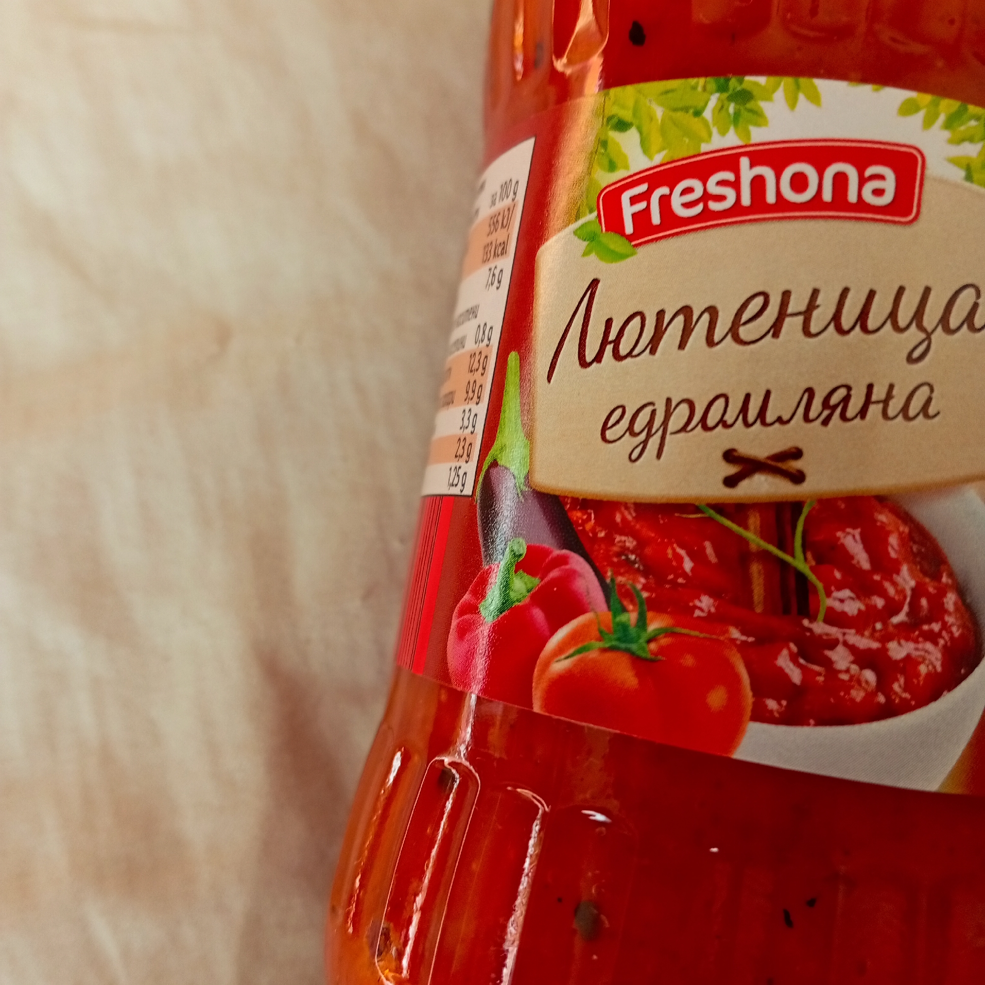 The scene is an indoor setting in Harmanli, Bulgaria. The primary subject is a transparent plastic bottle of "Freshona Lyutenitsa Edraliyana," containing a coarse, reddish-orange vegetable spread. The bottle is positioned on a light beige, textured surface. The product label features the brand "Freshona" in red text, above the Cyrillic inscription "Лютенuца едралияна," which translates to "Coarse Lyutenitsa." Graphical elements on the label illustrate ingredients, including a purple eggplant, a red bell pepper, and two red tomatoes, alongside a white bowl containing the finished lyutenitsa with visible vegetable pieces. Partial nutritional information is visible on the left side of the label, displaying values such as 556 kJ and 133 kcal per 100g. The bottle itself has vertical indentations and contains the chunky product, with small dark particles interspersed within the relish. No individuals, actions, or direct interactions are observable.