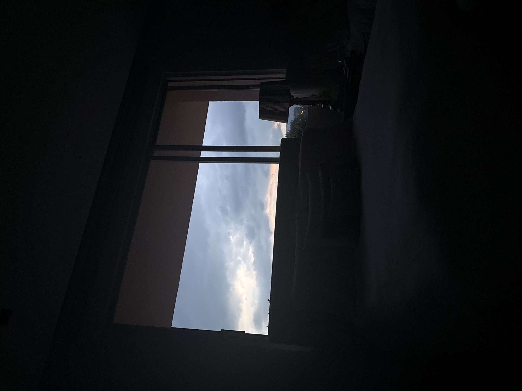 The image appears to be taken from inside a darkened room, looking out through a window towards a cloudy sky. The light from outside illuminates the clouds and the window frame, creating a contrast with the darkness of the room. A lamp and a partial view of furniture are visible within the room. The sky shows hues of blue, grey, and hints of pink, suggesting either sunrise or sunset. It is not possible to determine the location as Mexico City, Mexico, from the given image alone.