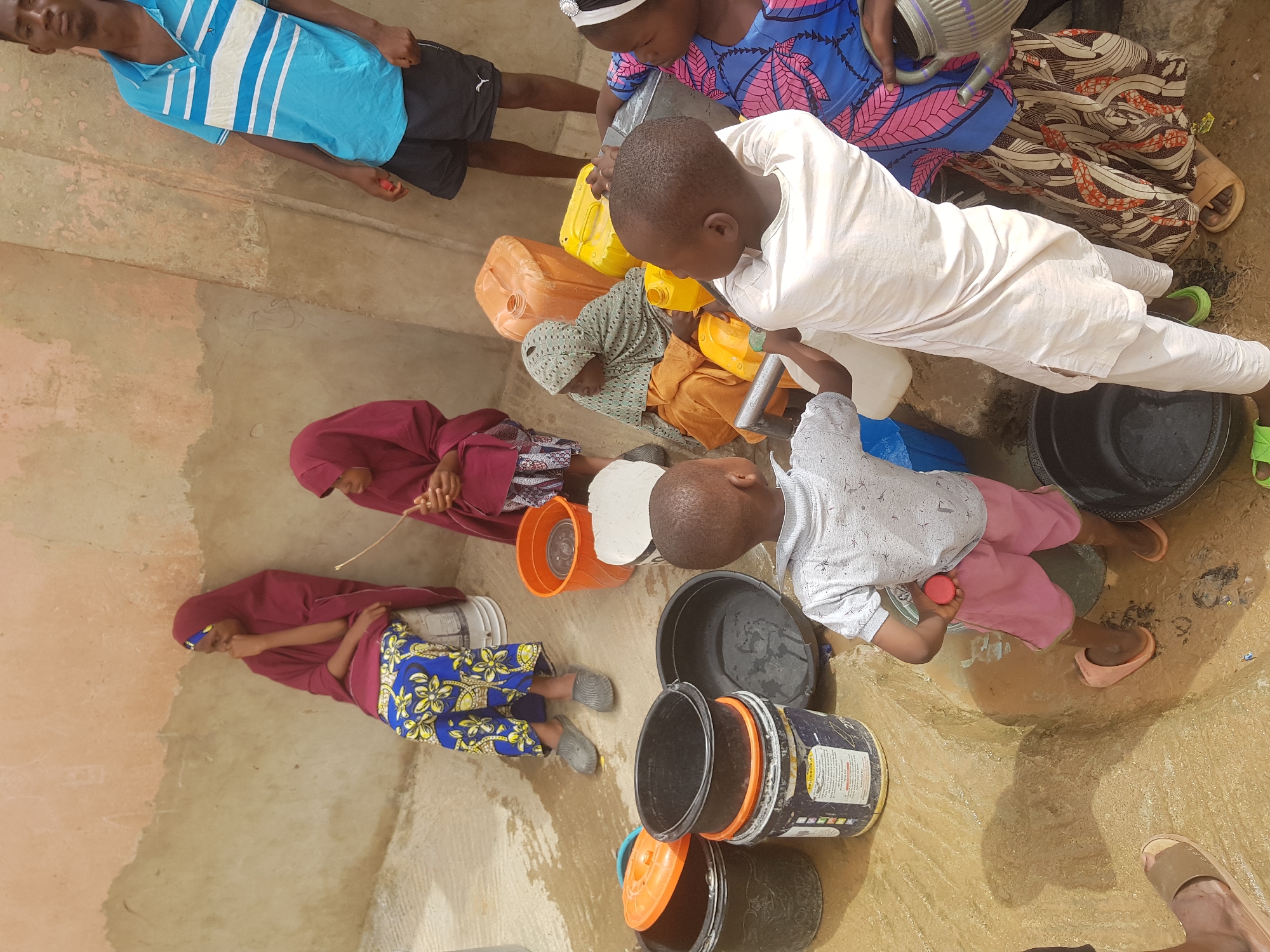 **People:** A group of children and adults, approximately seven individuals, are depicted. Some wear traditional clothing, including headscarves and patterned fabrics. One individual wears a blue and white striped shirt.
**Subjects:** The focus is on the process of collecting water.
**Objects:** Several containers are present, including buckets, plastic jerrycans (yellow and blue), and metal water vessels.
**Actions:** The individuals are gathered around a water source, filling containers. Some appear to be pouring water.
**Interactions:** People seem to be working together to collect water.
**Environment:** The scene appears to be a communal water point in Dutsinma A, Nigeria. The ground is wet and muddy, indicating recent water spillage. The backdrop consists of a simple structure with a partially painted wall.