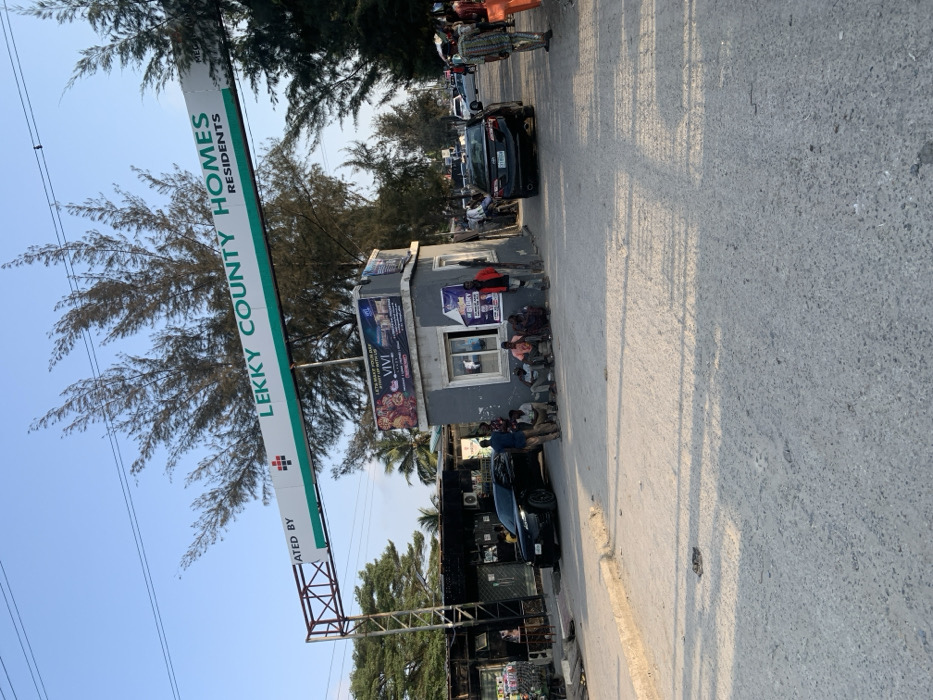 The scene depicts the main entrance to Lekky County Homes, located in Ikota, Lagos, Nigeria. A prominent metal archway spans a paved road, displaying a white and green sign with "LEKKY COUNTY HOMES RESIDENTS" and "CREATED BY" alongside a red and white logo. To the right of the entrance, a small, grey concrete security booth with windows and various advertisements affixed to its exterior is visible. Approximately seven to ten adult males are gathered around this booth; several are seated on the ground or a low ledge, while others stand nearby.
On the left side of the entrance, behind a parked dark-colored SUV, several small commercial structures or stalls with corrugated metal roofs are present. A black sedan is visible further down the road to the right, positioned as if entering or exiting the estate. The sky is clear blue, and tall, slender evergreen trees line both sides of the road and extend into the background. Overhead power lines are visible against the sky. The foreground consists of a grey, textured paved road surface, with concrete curbs defining its edges. Natural sunlight illuminates the scene, casting distinct shadows.