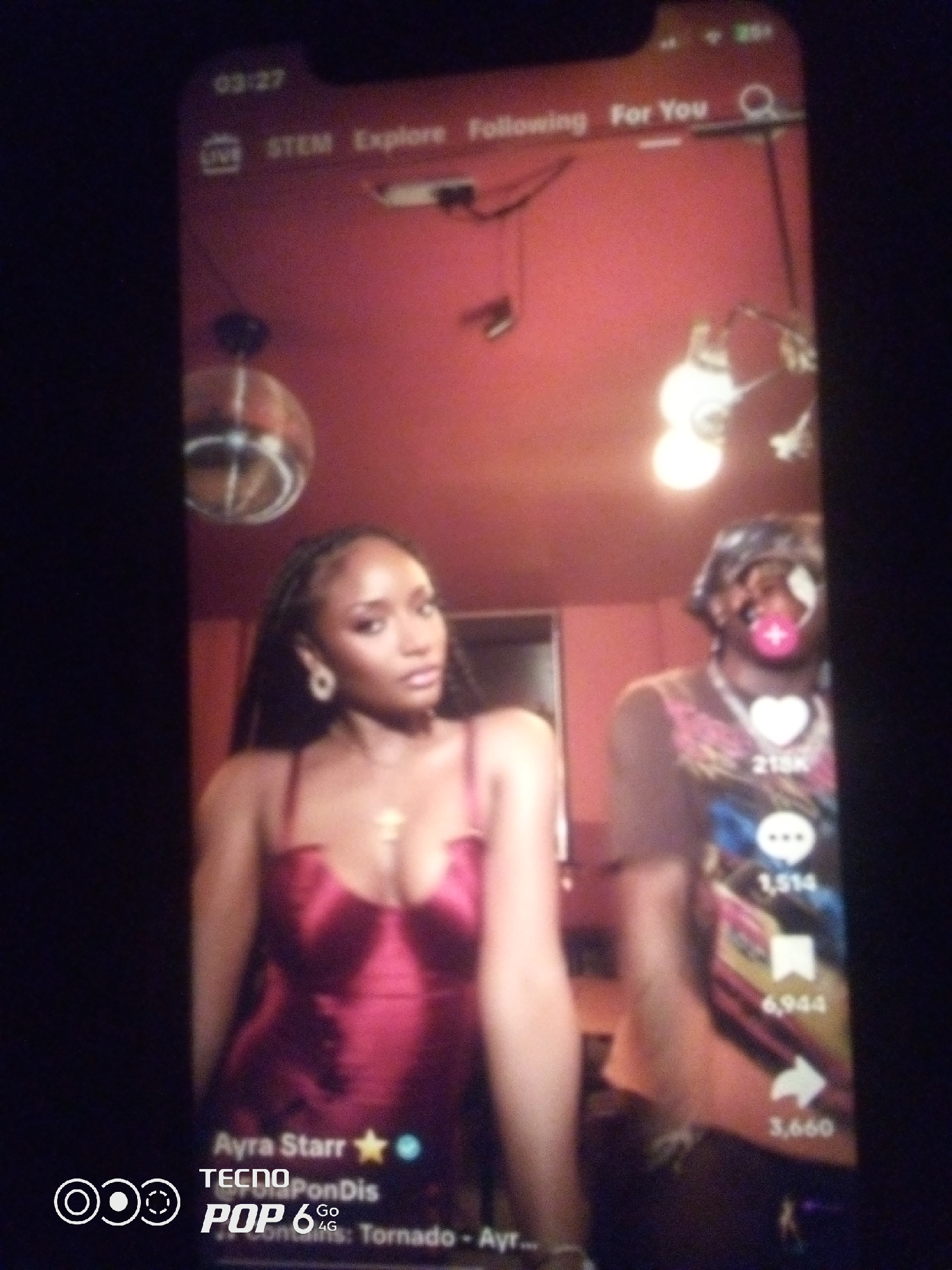 The image is a blurry, low-resolution photograph of a smartphone screen displaying a video. The video appears to be a social media clip, likely from a platform like TikTok, given the icons and text overlays common to such applications.
In the foreground, two individuals are visible. The person on the left is a woman with dark hair styled in braids, wearing a shimmering red satin dress and gold earrings. She is looking towards the camera with a neutral expression. The person on the right is wearing a bandana or headscarf and appears to have face paint or makeup on. They are positioned slightly behind and to the right of the woman.
The background of the video shows a reddish-brown wall and ceiling. There are several light fixtures visible, some appearing as glowing orbs. A disco ball is also hanging from the ceiling.
On the screen overlay, there are various UI elements typical of a video sharing app. These include:
* A timestamp at the top left reading "03:27".
* Navigation options like "LIVE", "STEM", "Explore", "Following", and "For You".
* A search icon.
* Interaction icons on the right side of the screen, including a heart icon with "215K" next to it, a speech bubble icon with "1,514" below it, a bookmark icon with "6,944" below that, and a share icon with "3,660" at the bottom.
* Text at the bottom of the screen which includes "Ayra Starr ★", "TECNO POP 6 4G", and further text that is partially obscured and illegible, but appears to be a caption or title related to the video.
The overall scene suggests a recording taking place indoors, possibly in a studio or a decorated room, with a focus on the two individuals and the video content displayed on the phone. There is no visual information in the image that can be used to determine its location in the city of Howayi, Nigeria.