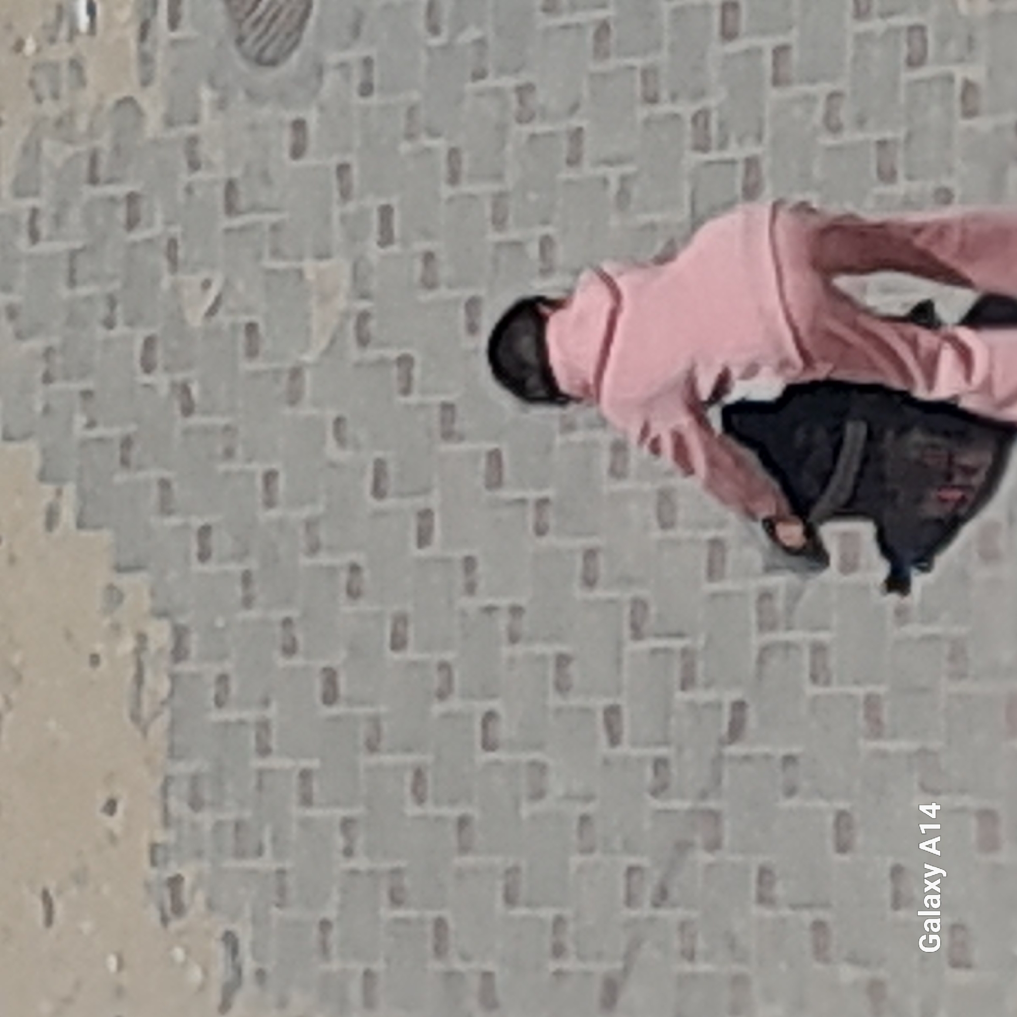 The image captures an outdoor scene featuring a single person walking on a patterned paved surface. The individual, who appears to be a child or a young person, is seen from behind, moving away from the camera with their head slightly turned to the left. They are wearing a light pink long-sleeved top and matching pants, possibly part of a tracksuit. A dark-colored backpack is visible on their back.
The ground is covered with light gray paving stones arranged in an intricate, interlocking pattern. On the left side of the frame, there is a lighter, irregular area, possibly dirt or a different ground cover, adjacent to the paved section. The lighting suggests it is daytime, and the weather appears clear and dry. No strong shadows are present, making it difficult to determine the exact time of day. The person's expression is not visible.
In the bottom right corner of the image, the text "Galaxy A14" is visible vertically, indicating the camera or device used to capture the photograph.