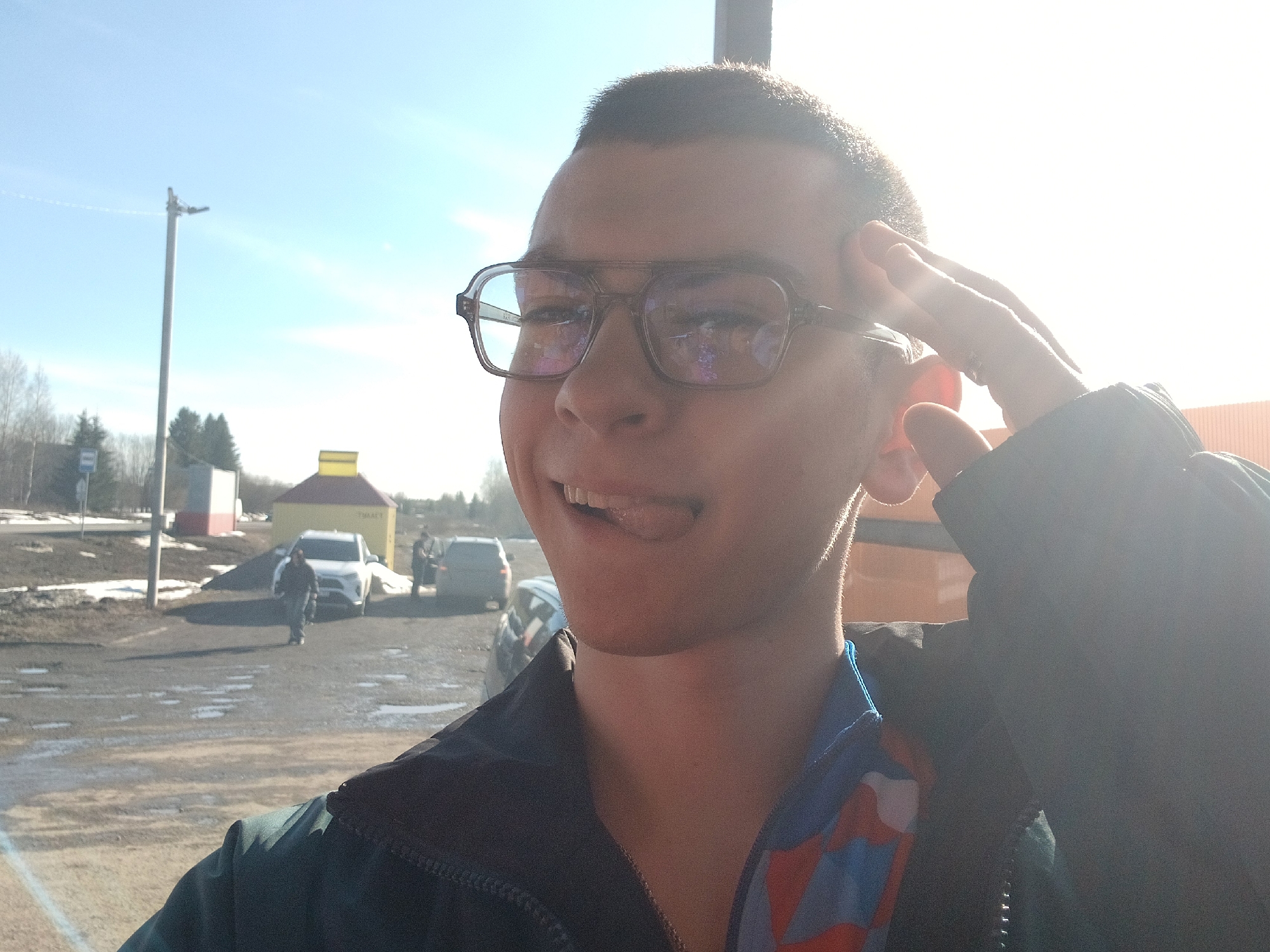 A medium, frontal shot depicts a person with short, dark hair and eyeglasses, sticking out their tongue. The person is wearing a dark jacket with a blue and red underlayer visible at the neck.
In the background, a parking area is visible with several cars, including a white SUV. In the distance, there's a yellow structure with a sign in Russian, located near Gory, Russia. A person is walking near the SUV. A utility pole stands to the left, and remnants of snow are visible on the ground. The sunlight is bright.