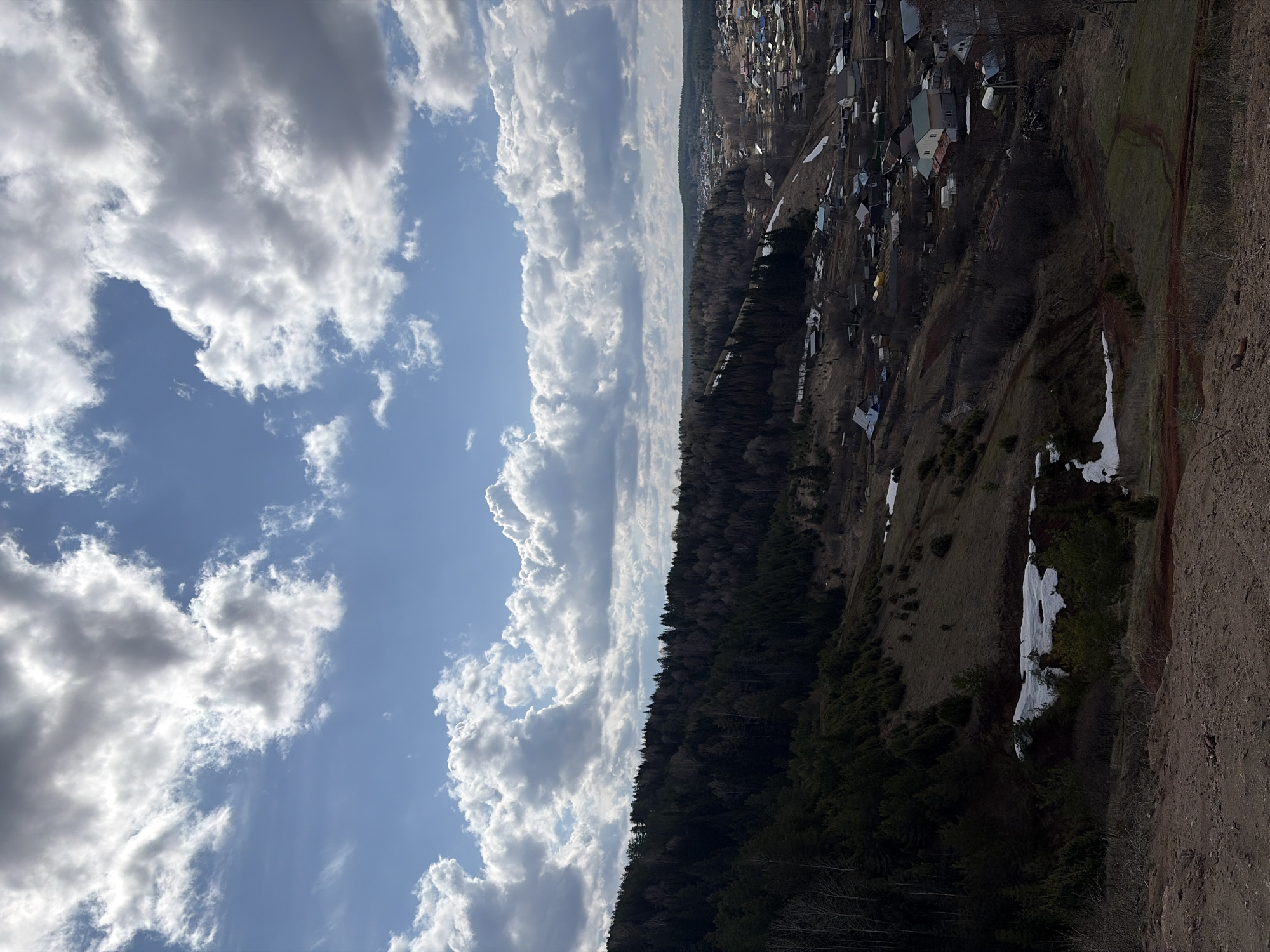 The image shows a landscape view from a high vantage point in or near Ольховка, Russia. The foreground consists of a dry, rocky hillside, with some patches of melting snow. Beyond the hillside is a dense forest, and beyond that, a cluster of houses and buildings indicates the edge of a village. The sky occupies a significant portion of the frame, featuring a mix of blue sky and scattered cumulus clouds.