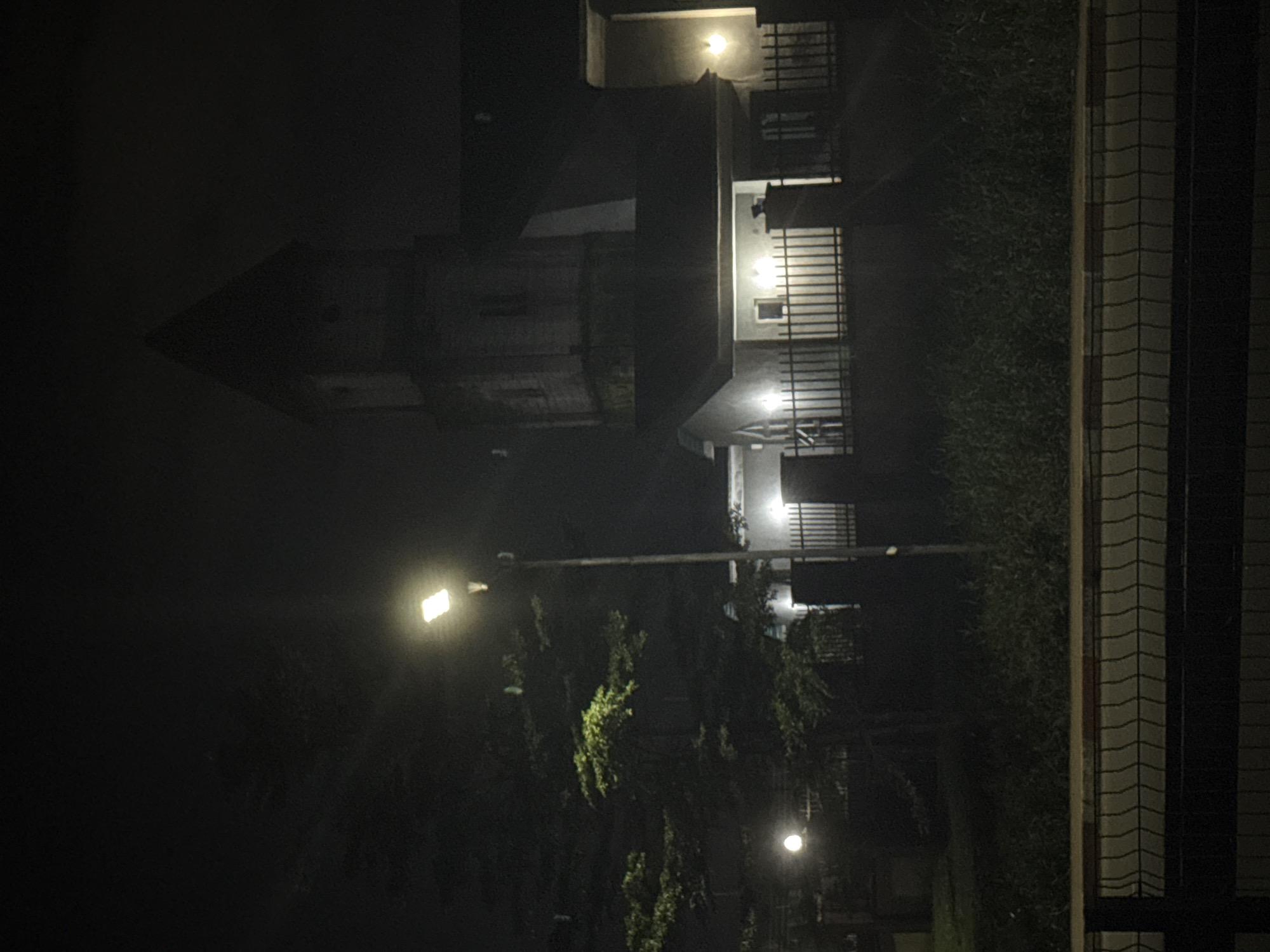 This image depicts a nighttime scene in Uyo, Nigeria. In the background, a building with a distinctive tall, pointed tower, reminiscent of a church or observatory, is illuminated by artificial lights. The building appears to be constructed of concrete and has a dark, pitched roof. A fence separates the building from the foreground. To the left of the building, a streetlamp with a bright, rectangular light fixture casts a glow on the surrounding area, illuminating a tree with dense foliage. Further to the left, another smaller, round light source is visible. The foreground is dominated by a metal fence with a zig-zag pattern. The ground is covered in grass and vegetation. The overall atmosphere is dark and somewhat mysterious, suggesting a quiet, possibly residential or institutional area.