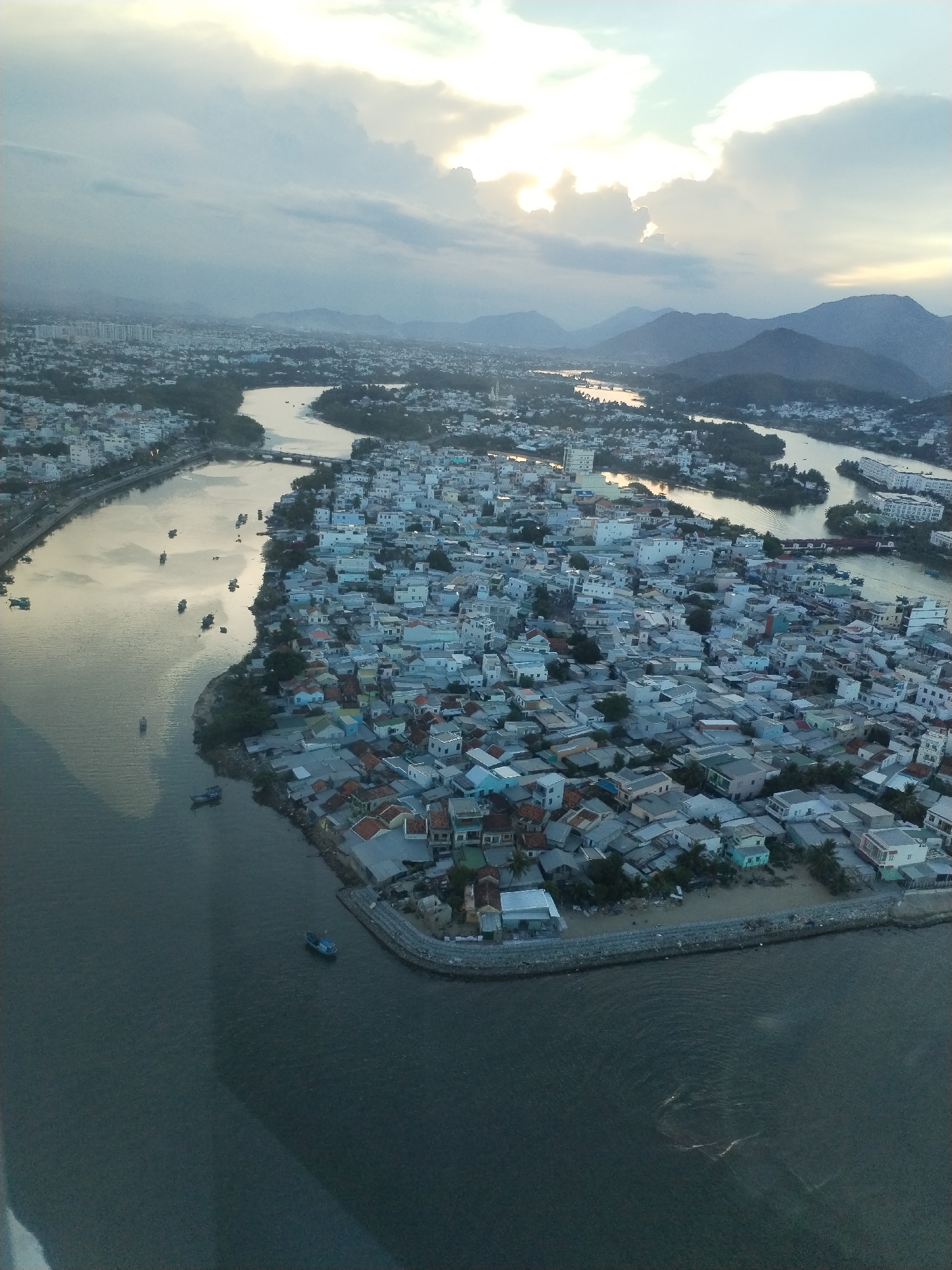 Aerial view of Bac Nha Trang Ward, Vietnam, at dusk. Predominantly low-rise buildings with a mix of red tile and other roofing materials. The architecture is dense, and tightly packed. Rivers and waterways meander throughout the landscape, reflecting the setting sun. Several small boats are present on the water. Distant hills and mountains are visible on the horizon beneath a cloudy sky.