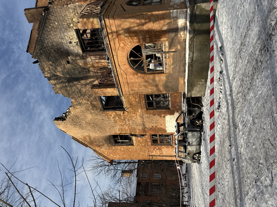 This image depicts an urban scene in Kyiv, Ukraine, showcasing the aftermath of extensive damage to a building and vehicle in a winter setting. The primary subject is a multi-story structure, constructed of orange-brown brick, exhibiting widespread charring and soot deposition across its facade. The upper left portion of the building displays severe structural compromise, with a jagged, irregular roofline and wall. All visible windows are shattered, lacking glass, with frames either missing or heavily burnt. A metal balcony railing on the upper right side is present but appears damaged. In the foreground, positioned on snow-covered ground in front of the building, is the fire-gutted chassis of a small vehicle, resembling an SUV or hatchback, reduced to charred metal components. A red and white striped barrier tape horizontally spans the lower portion of the frame, indicating a secured area. The ground is covered with dirty snow, and bare tree branches are visible against a partly cloudy blue sky. No individuals or active operations are depicted.