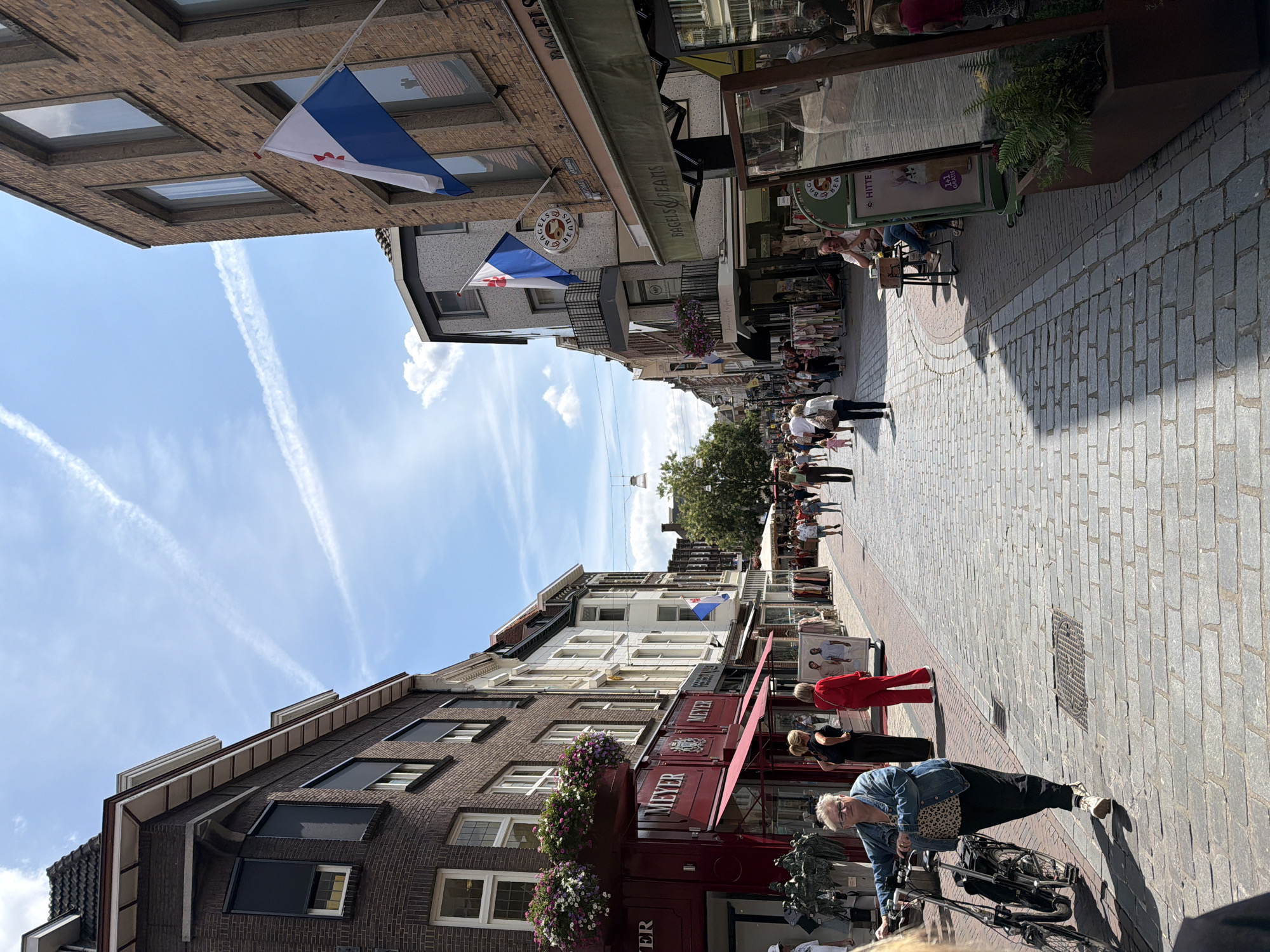 The image depicts a street scene in the city of Roermond, Netherlands. The street is paved with cobblestones and lined with buildings. On the left, a dark brick building with large windows and awnings houses a store called "MEYER". Flower boxes filled with colorful flowers are attached to the building. Several people are walking along the street.
Further down the street, on the right side, a brick building has a flag flying from it. Next to it, a store called "BAGELS & BEANS" is visible, with awnings extending outwards. More people are seen walking and sitting at tables outside the store. The sky is blue with scattered white clouds and contrails from airplanes. The overall scene suggests a busy urban environment on a sunny day.