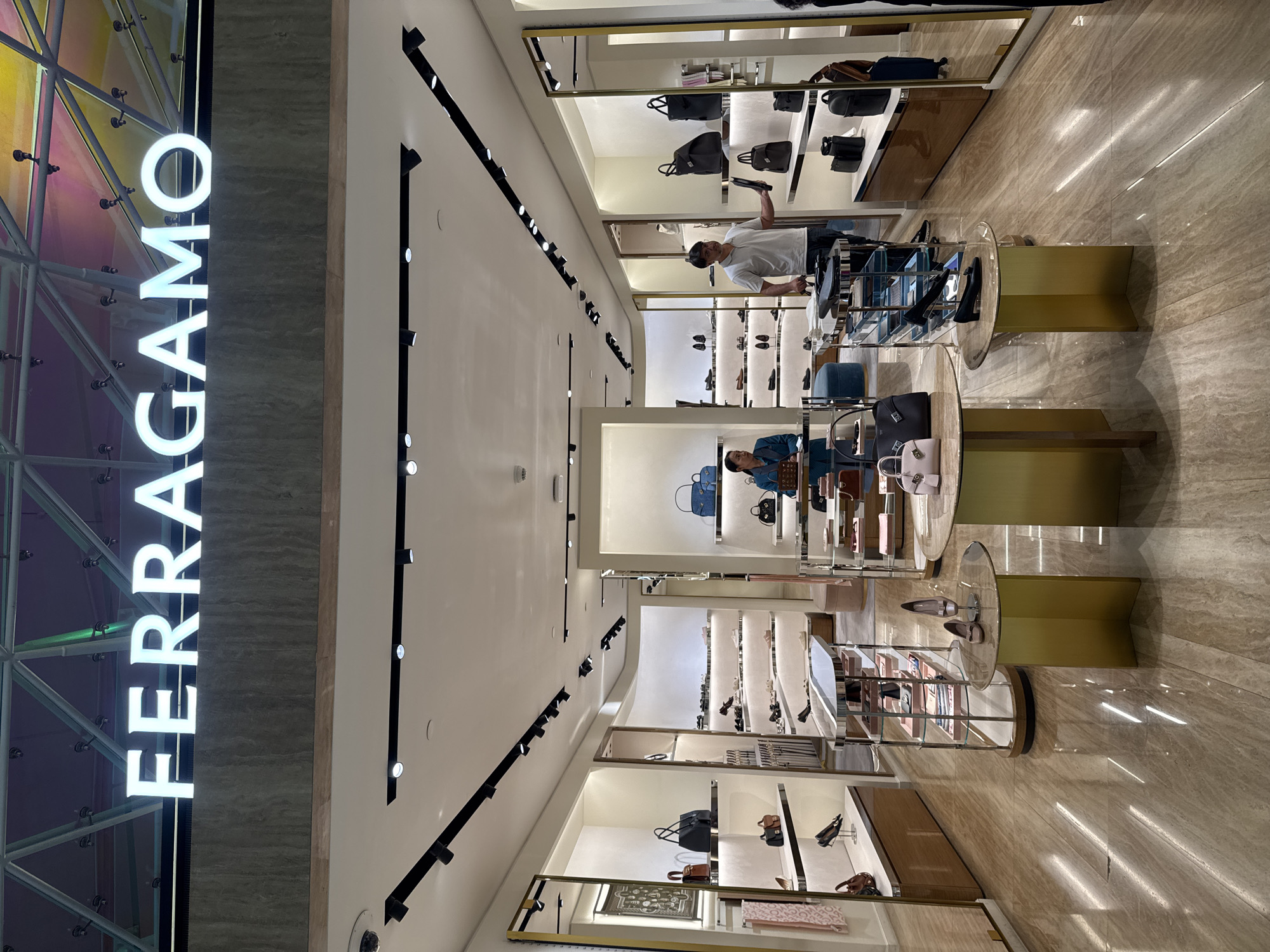 The image shows the interior of a Ferragamo store, likely located in a shopping mall. The store is well-lit with bright overhead lighting and display lights illuminating the merchandise. The floor is made of polished marble tiles with a reflective surface.
In the foreground, there are two round display tables with gold-colored bases. The left table holds a light pink handbag, and the right table displays a pair of beige high-heeled shoes. A narrow display rack with multiple shelves containing handbags is visible behind the right table.
Further into the store, there are numerous shelves stocked with shoes and handbags. A woman in a teal blazer is standing behind a counter on the left side of the image, and a man in a light grey polo shirt is standing in the middle of the store, holding a black shoe.
The store's name, "FERRAGAMO," is prominently displayed in large white illuminated letters above the entrance. The background shows the colorful, iridescent ceiling of what appears to be a mall.
The location is the city of Nong Prue Subdistrict, Thailand.
