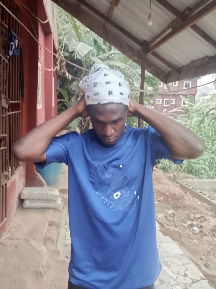 A young, dark-skinned man in a blue t-shirt and a patterned white bandana stands outside, holding his hands to the back of his head. He is facing forward with his eyes looking down. Behind him, there is a maroon building with a barred window, a wooden roof structure, and foliage. In the background, other buildings and more greenery are visible. The ground is paved with stones.