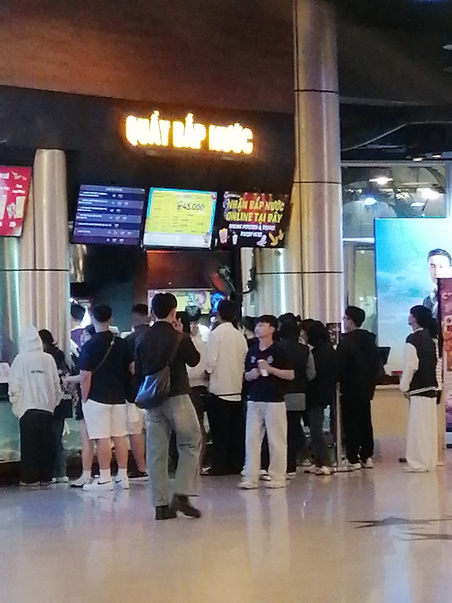 The image depicts a group of people standing in front of a brightly lit counter labeled "QUAY BẮP NƯỚC" (Popcorn and Drink Counter) in what appears to be a cinema lobby. The signage above the counter is in Vietnamese and features a glowing orange text. To the left of the counter, a digital display shows movie showtimes, and to the right, another screen advertises "NHẬN BẮP NƯỚC ONLINE TẠI ĐÂY" (Pick up popcorn and drinks here online) with prices.
Several individuals are visible, some facing the counter, others looking towards the right. They are dressed casually, with a mix of t-shirts, hoodies, and pants. One person in the foreground is wearing a black t-shirt and jeans, with a bag slung across their shoulder. Another person on the left is wearing a white hoodie. The environment appears to be an indoor public space with a polished floor reflecting the lights. The overall impression is a busy scene at a ticket or concession stand, likely within a cinema or entertainment venue in Da Lat, Vietnam.