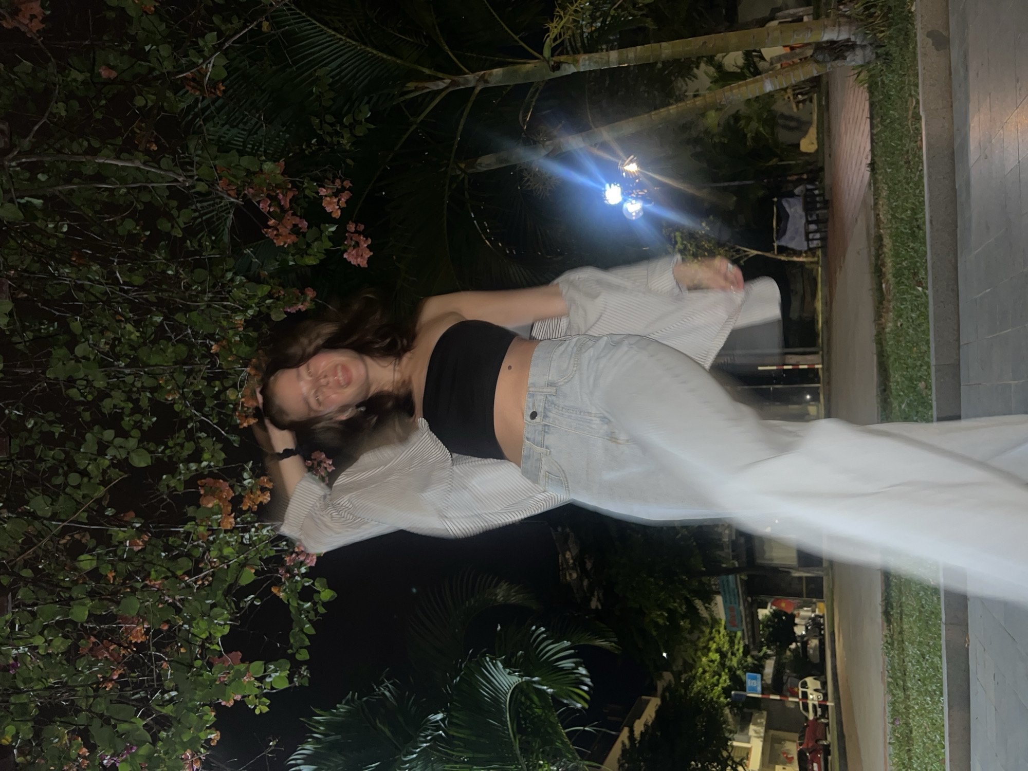 A woman is standing outside in Nam Nha Trang Ward, Vietnam. She wears a black tube top, light blue jeans, and a white striped shirt. She smiles, and her right arm is raised. The environment includes trees with orange flowers, palm trees, and street lights. A sidewalk and buildings are visible in the background.