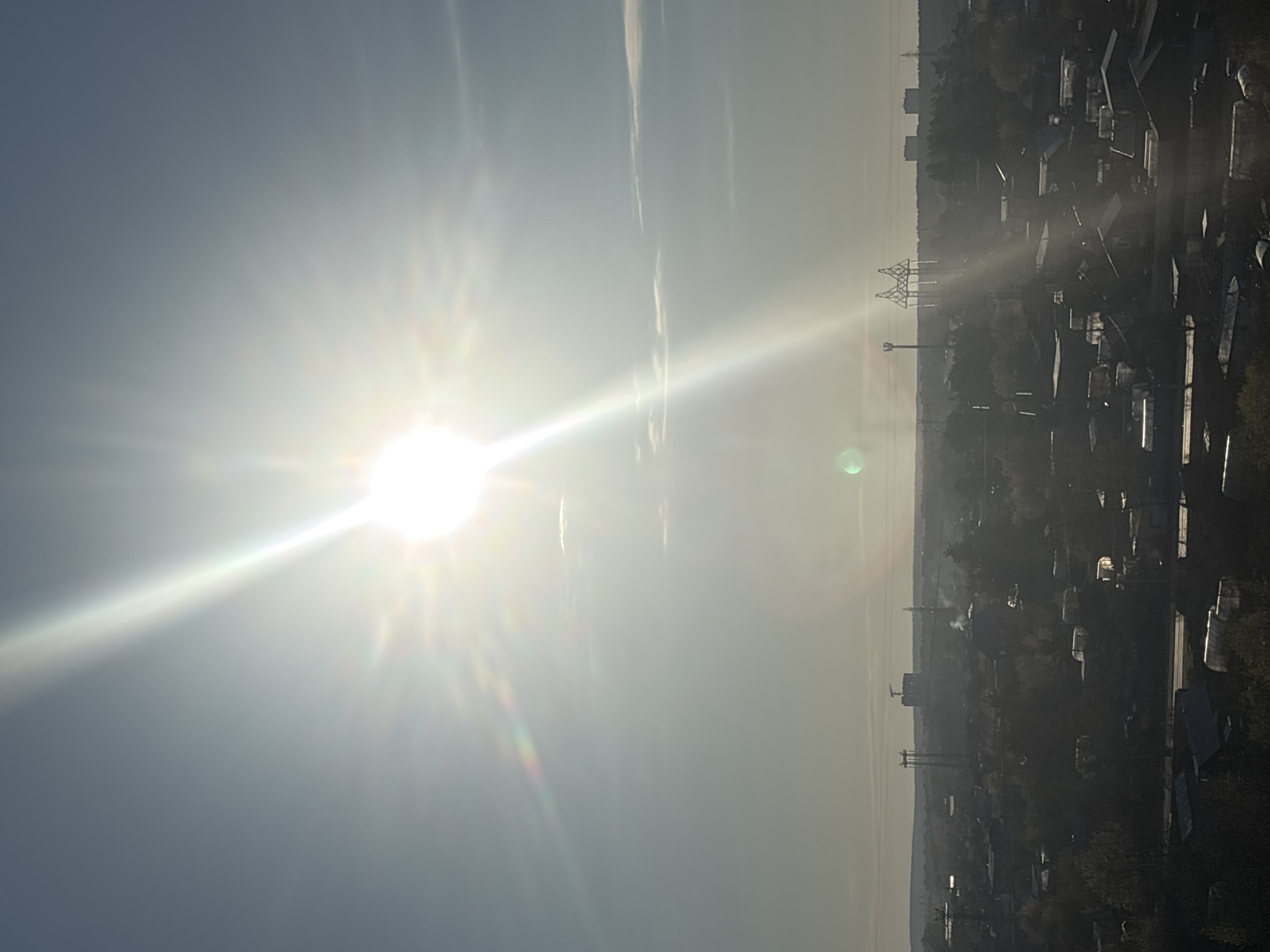 The image shows a hazy, high-angle view of a residential area in Chaikovsky, Russia, under a bright sun. The sun is positioned to the left of the frame, causing glare and partially obscuring the sky. In the distance, the cityscape is visible, with outlines of buildings and structures along the horizon. Power lines and towers are seen amidst the urban landscape. The foreground consists of rooftops of houses and trees, giving the impression of a dense residential area. The overall scene is washed in sunlight, causing shadows and limiting visibility in some areas.