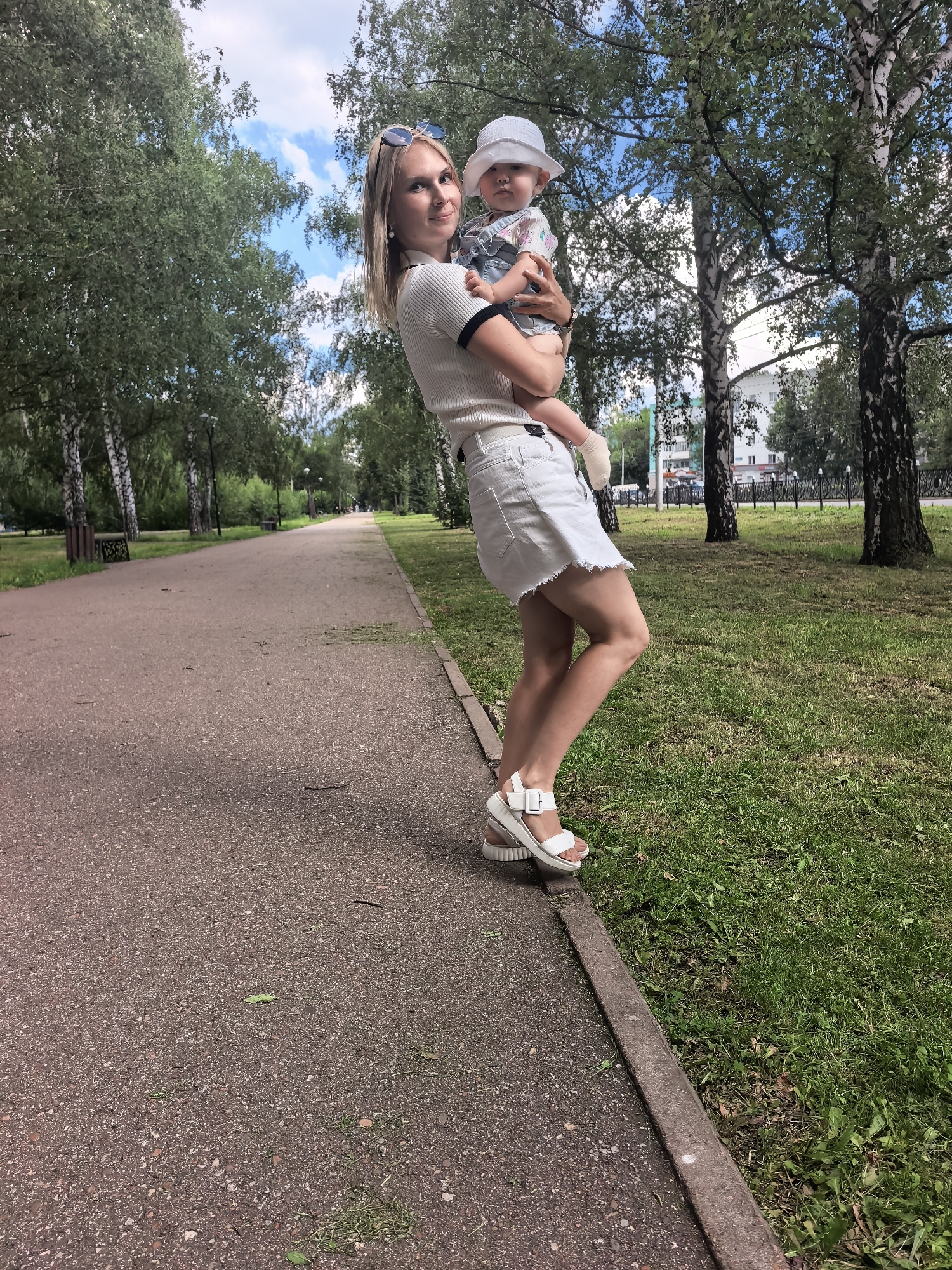 A blonde woman in a white top and skirt, wearing white sandals and sunglasses, holds a baby in a white hat and denim overalls. They are standing on a paved path in a park lined with trees. The woman is looking at the camera, and the baby is looking slightly to the side. The park is in the city of Sterlitamak, Russia, indicated by the birch trees and the architecture in the background.