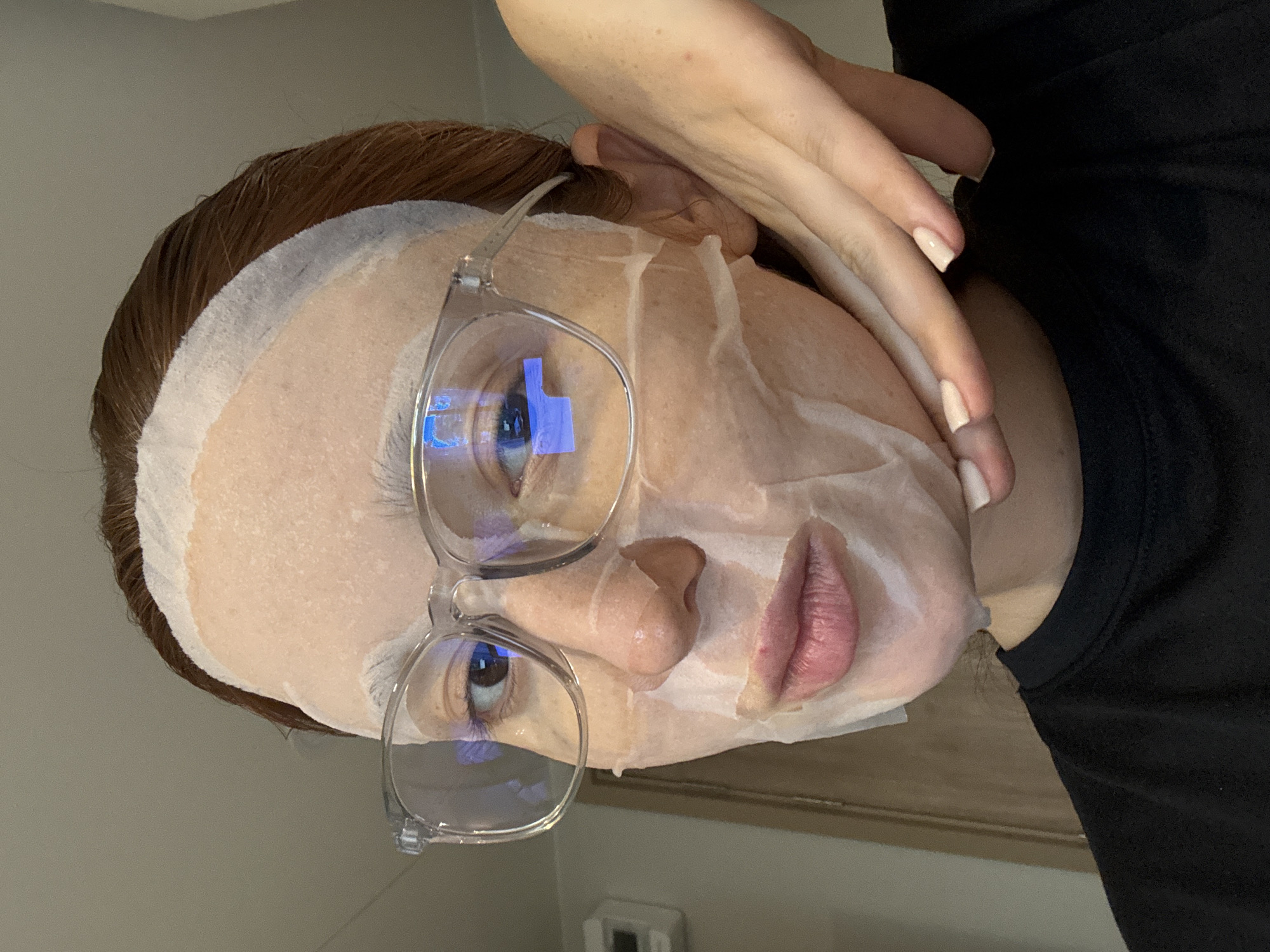 A person wearing a facial mask and glasses is looking at the camera. The person has light skin, auburn hair, and is wearing a black shirt. Their left hand is raised to their face. The background appears to be an indoor setting, possibly a bathroom. It is impossible to ascertain the location is Choeng Thale, Thailand.