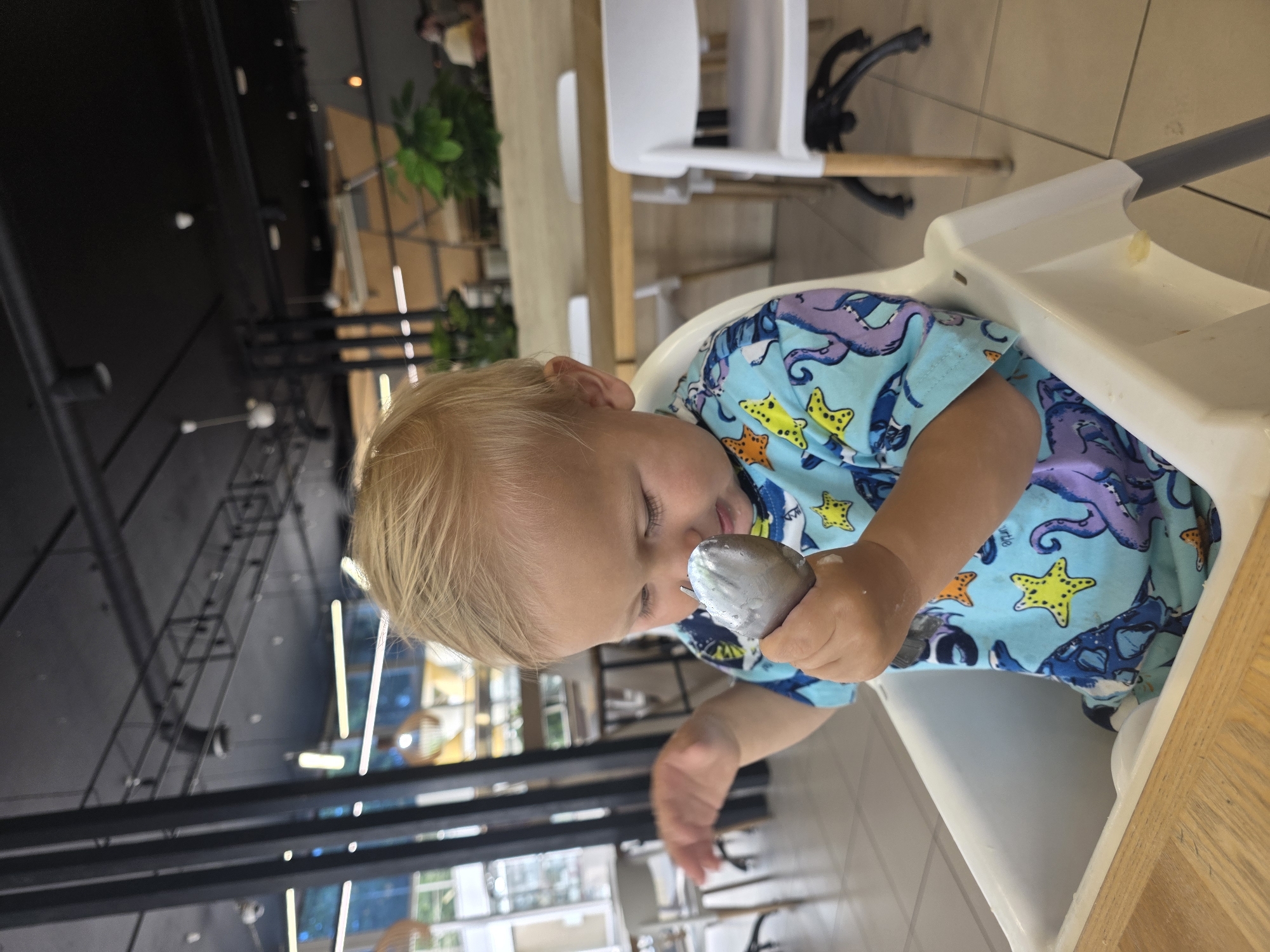 A young child with blonde hair is sitting in a white high chair at a wooden table. The child is wearing a blue shirt with an octopus and starfish pattern. The child is holding a silver spoon, looking down at it with closed eyes. It appears the child might be eating or playing with the spoon. The background shows a bright, modern interior with tables, chairs, and plants. The overall lighting suggests it is daytime. The scene is likely a restaurant or cafe.
