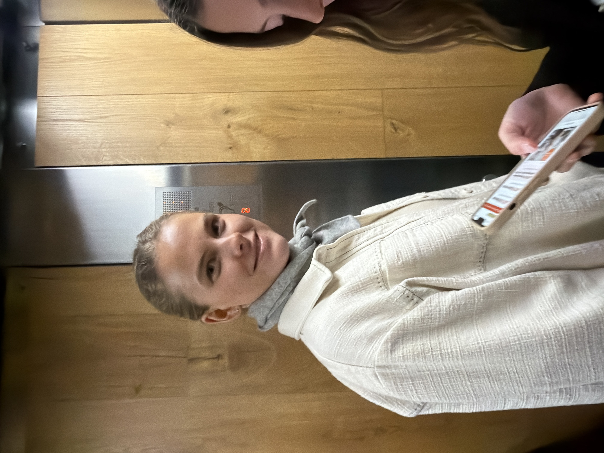 The image depicts two individuals inside an elevator. One person, a fair-skinned female with blonde hair pulled back, is in focus, smiling slightly towards the camera. She wears a white textured jacket over a grey hooded sweatshirt. She appears to be positioned near the elevator door, with a stainless steel panel visible behind her. The elevator walls feature a wood-grain pattern. The second person, also fair-skinned, has long brown hair and is partially visible at the top of the frame. A hand holding a smartphone with an illuminated screen is also visible. Without further details, it is impossible to determine with certainty if the scene is located in London, UK.
