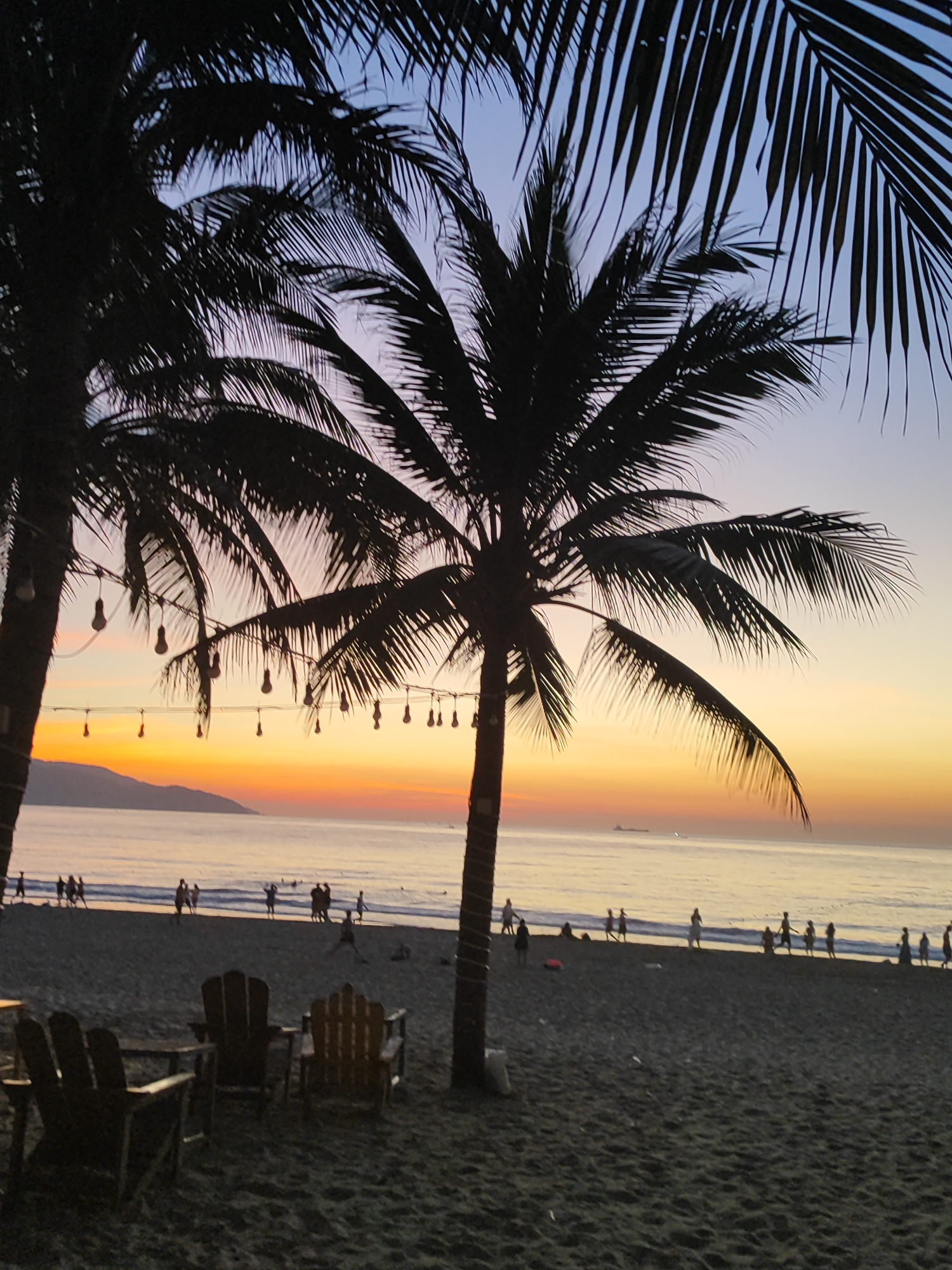 The image depicts a twilight scene at My Khe Beach in Da Nang, Vietnam. Silhouetted palm trees frame a view of the ocean and the sandy beach. The sky displays a gradient of colors, transitioning from a pale blue at the top to hues of orange and yellow near the horizon, indicative of sunset. String lights with exposed bulbs hang from the palm trees in the foreground.
On the beach, several people are visible, mostly in silhouette. Some are walking along the shoreline, while others appear to be engaged in activities such as wading in the shallow water or standing in groups. A few beach chairs are set up on the sand, suggesting a relaxed atmosphere. In the distance, a ship can be seen on the water, and a mountainous coastline forms the backdrop. The sand is dark, likely due to the low light conditions.