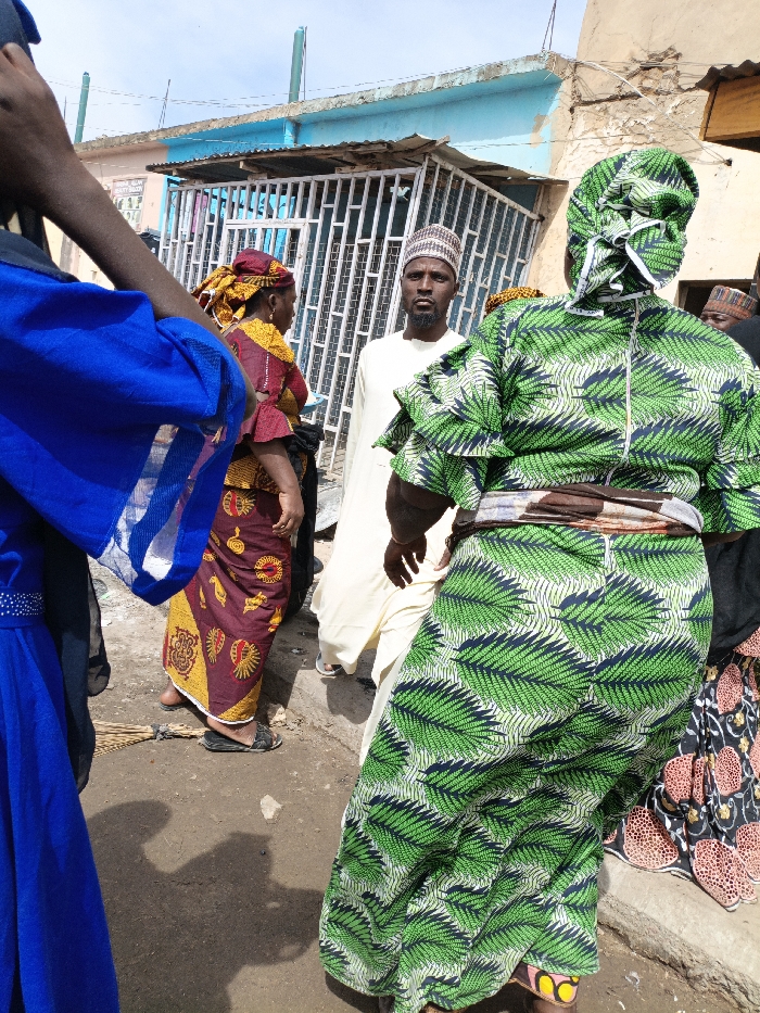 The image depicts an outdoor street scene in Gama, Nigeria, with multiple individuals. In the foreground, partially visible on the left, is a person wearing a blue garment with white trim. On the right, a woman in a green and dark blue leaf-patterned dress is seen from behind, with a brown fabric sash tied around her waist. In the mid-ground, a bearded man wearing a light-colored robe and a patterned cap (kufi) looks directly at the viewer. To his left, a woman in a maroon, yellow, and gold patterned dress and a matching headwrap is partially obscured, facing away from the camera. Another individual with a patterned cap is partially visible in the background to the right. The ground is unpaved dirt, showing minor debris, including a bundle of sticks near the left mid-ground woman's feet and a small light-colored stone in the center. In the background, a light blue building with a corrugated metal overhang and a white metal security gate is visible. Two green poles extend from the building's roof, and a faded sign with partial text is present on its wall. The sky above is light blue.