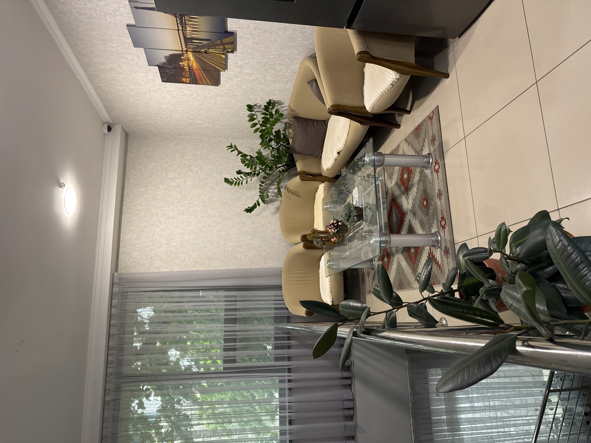 This media file captures an interior scene, likely a living room or waiting area, in Bishkek City, Kyrgyzstan, presented in a landscape orientation after correction from the original rotated view. The space is furnished with two beige upholstered armchairs featuring wooden armrests and cream-colored textured panels, arranged facing each other with a rectangular glass coffee table between them. Underneath the table, a geometric patterned rug in red, grey, and white adds a touch of color to the light-colored tiled floor.
Behind the armchairs, a tall, vibrant green potted plant stands. Another large, dark-leafed potted plant is positioned in the foreground on the right, partially obscured by a silver railing. On the back wall, a multi-panel artwork depicts a night scene of a bridge or road illuminated by the streaks of car lights. The walls are white with a subtle textured finish, and a similar texture is visible on the ceiling.
A large window on the left is fitted with sheer horizontal blinds, allowing natural light to filter into the room, with green tree foliage visible outside, suggesting daytime. A white wall sconce provides additional lighting on the left, and a small white security camera is mounted high on the wall near the ceiling. A dark grey or black furniture piece, possibly a cabinet or refrigerator, is partially visible on the right. The room presents a clean, orderly, and functional environment. No individuals or visible text are present.