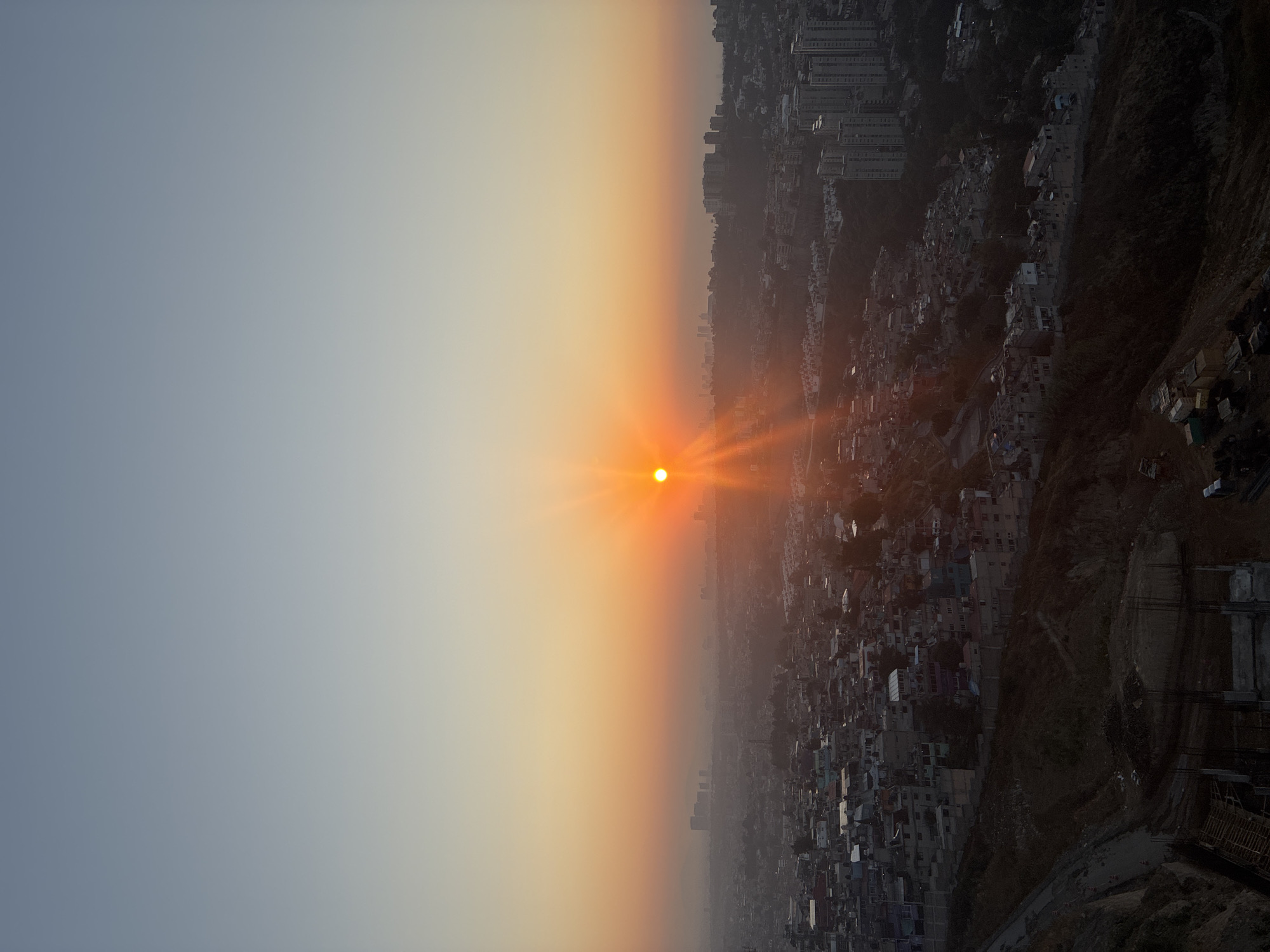 The image depicts a city, likely Huixquilucan de Degollado, Mexico, under a hazy, orange sunset. The sun, a bright orange orb, is positioned near the horizon, casting strong rays. A densely packed urban area with buildings of varying heights stretches out below the sun. The landscape fades into darker, hilly terrain on the right side of the frame, transitioning into a blue-grey sky. The image seems to have been taken from a high vantage point, overlooking the cityscape at dusk. There are no people visible in the image. The focus is on the urban environment and the sunset.