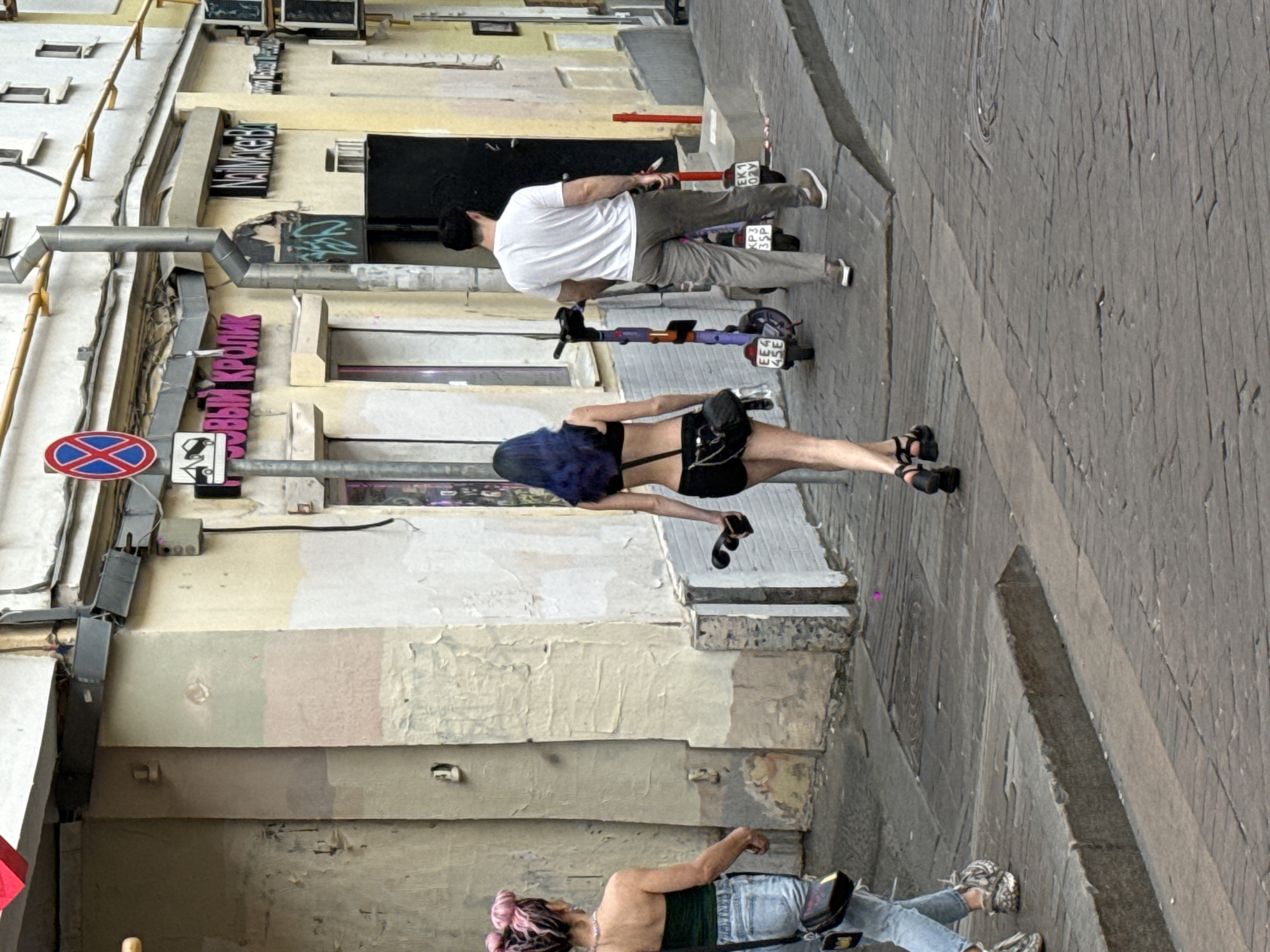 The image depicts a street in Moscow, Russia. Three people are present in the scene. One man, wearing a white shirt and khaki pants, is riding a scooter. A woman with blue hair is also riding a scooter. Another woman, wearing a green top and jeans, is walking and appears to be using a crutch or walking stick. Street signs and business names are visible in Russian. The buildings along the street show varied architectural styles with aged and textured facades. The sidewalk is made of stone tiles.