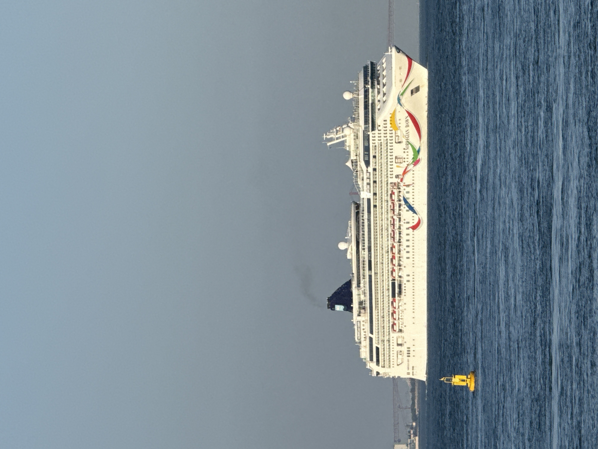 A large white cruise ship, the Norwegian Dawn, sails on a calm, deep blue ocean under a clear, pale blue sky. The ship features a distinctive rainbow design along its hull and the text "NORWEGIAN DAWN" is visible. A black plume of smoke emerges from the ship's funnel. In the foreground, a yellow buoy with a black top floats on the water. In the distance, a suspension bridge and the faint outline of a city, likely Oeiras, Portugal, are visible on the horizon. The overall scene depicts a serene day at sea.