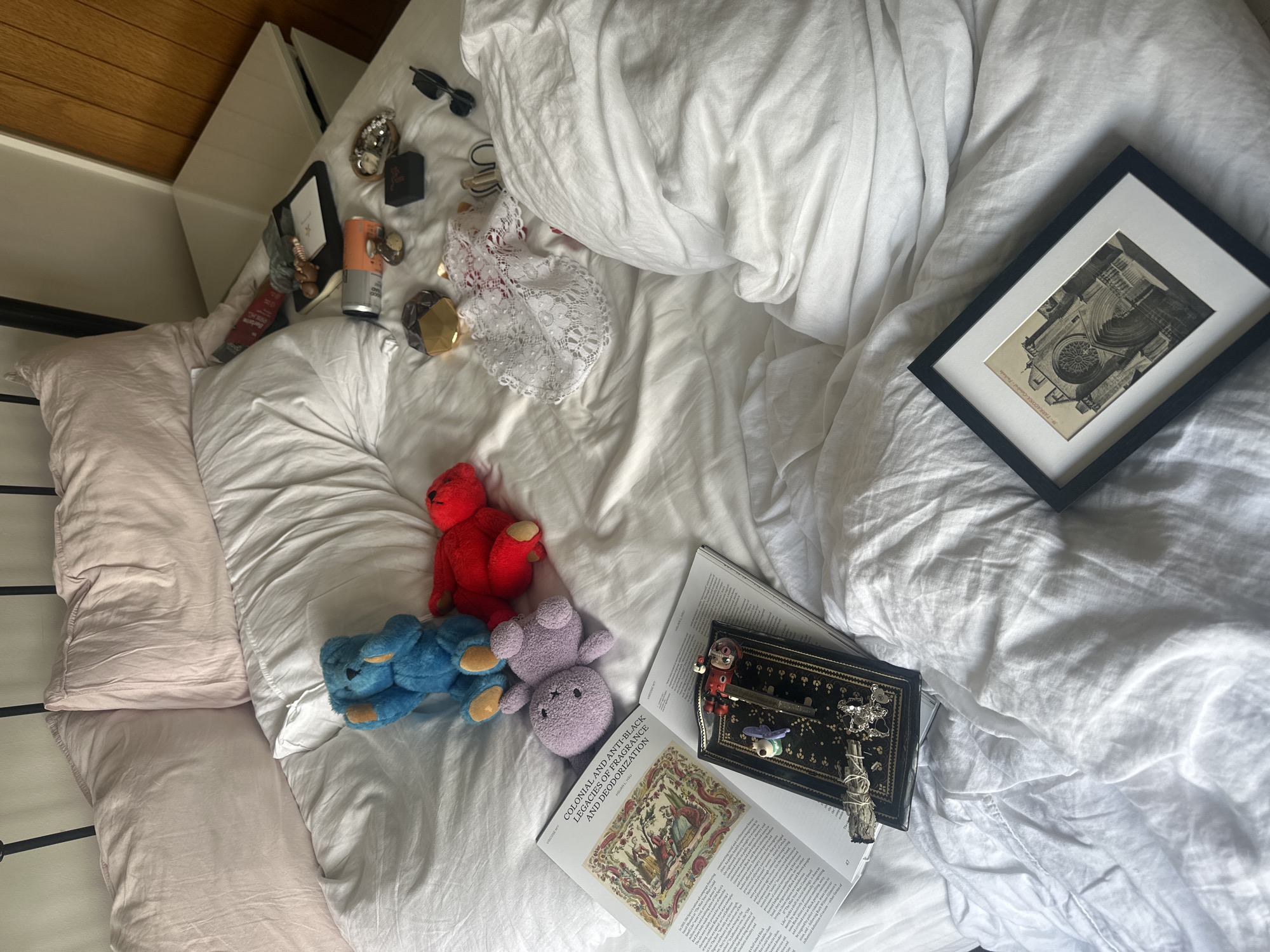 The image depicts a cluttered bedroom scene, specifically a bed with white linen duvet cover and two light pink pillows against a black metal headboard. To the right, a wooden wall and a white nightstand are visible.
On the bed, a blue plush teddy bear is seated with its hands near its face, beside a red plush teddy bear lying on its side, and a lavender plush teddy bear also lying on its side. An open magazine or journal titled "COLONIAL AND ANTI-BLACK LEGACIES OF FRAGRANCE AND DEODORIZATION" lies face-up, displaying text and an ornate illustration. A second open magazine is partially visible beneath it.
Resting on the open magazine is a black rectangular tray with gold decorative trim, holding various small items: a white skull charm, a purple butterfly ornament, a miniature metallic violin, a bundle of white sage, and a silver charm resembling an intricate animal.
Further to the right, towards the nightstand, are several personal items scattered on the bed and nightstand: a silver and orange can (possibly a beverage), a gold and clear glass perfume bottle, a small black box, a tray containing rings and other jewelry, a black electronic device, and a white lace garment with red accents underneath.
At the bottom right of the bed, a black-framed picture with a white mat displays a monochrome image of a cathedral facade, identifiable as "TARRAGONA Catedrale" from faint text on the original postcard within the frame.
The precise location within the city of Wealden, United Kingdom, cannot be determined from the visual information provided in the image.