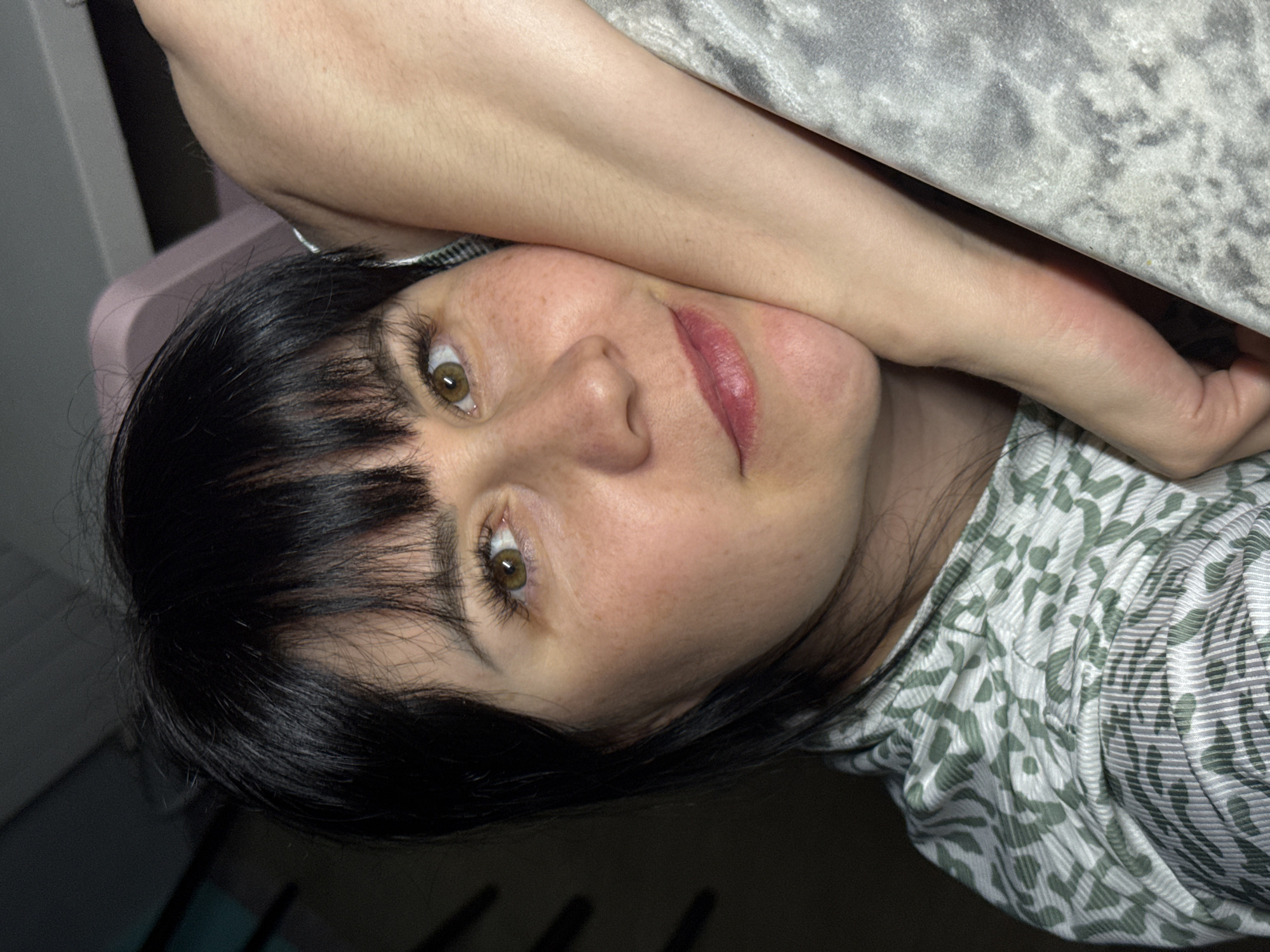 The image shows a woman with black hair and hazel eyes. She has fair skin with freckles and is wearing lipstick. Her left arm is raised and resting on a gray surface. She is wearing a patterned shirt. The background includes a pink object and a dark, undefined area. The image does not provide any information to determine that this photo was taken in Zav'yalovo, Russia.